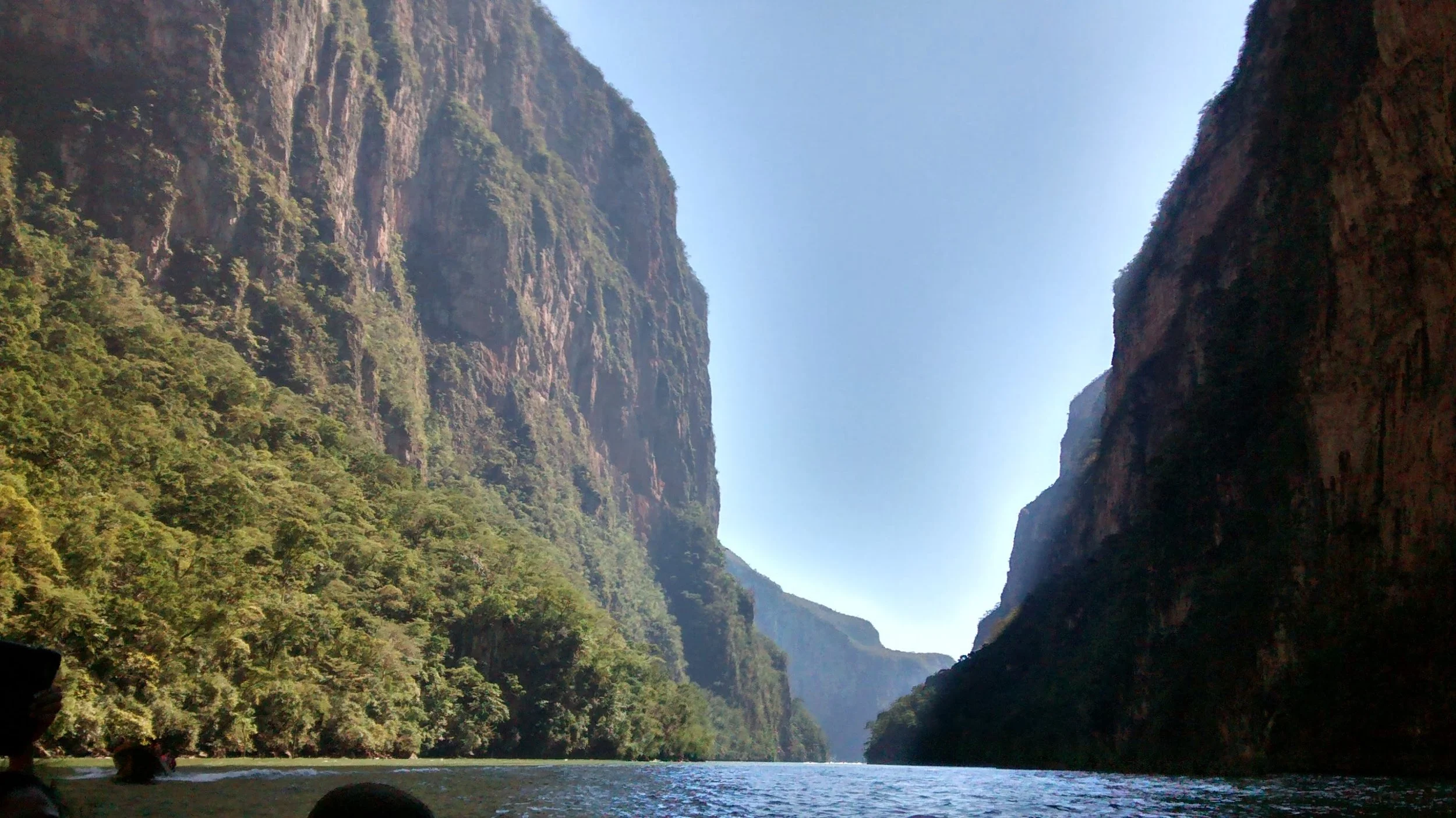 A river flowing between lush green mountains with steep cliffs on both sides under a clear blue sky.