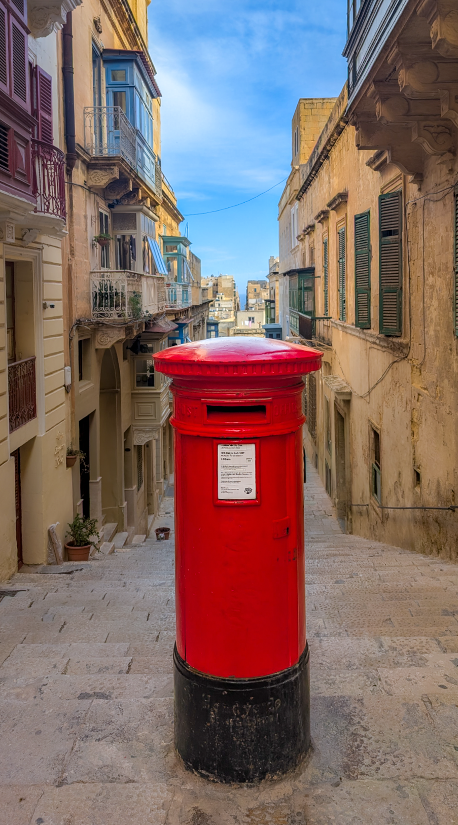 Red postal mailbox on a narrow European street with old buildings and blue sky in the background.