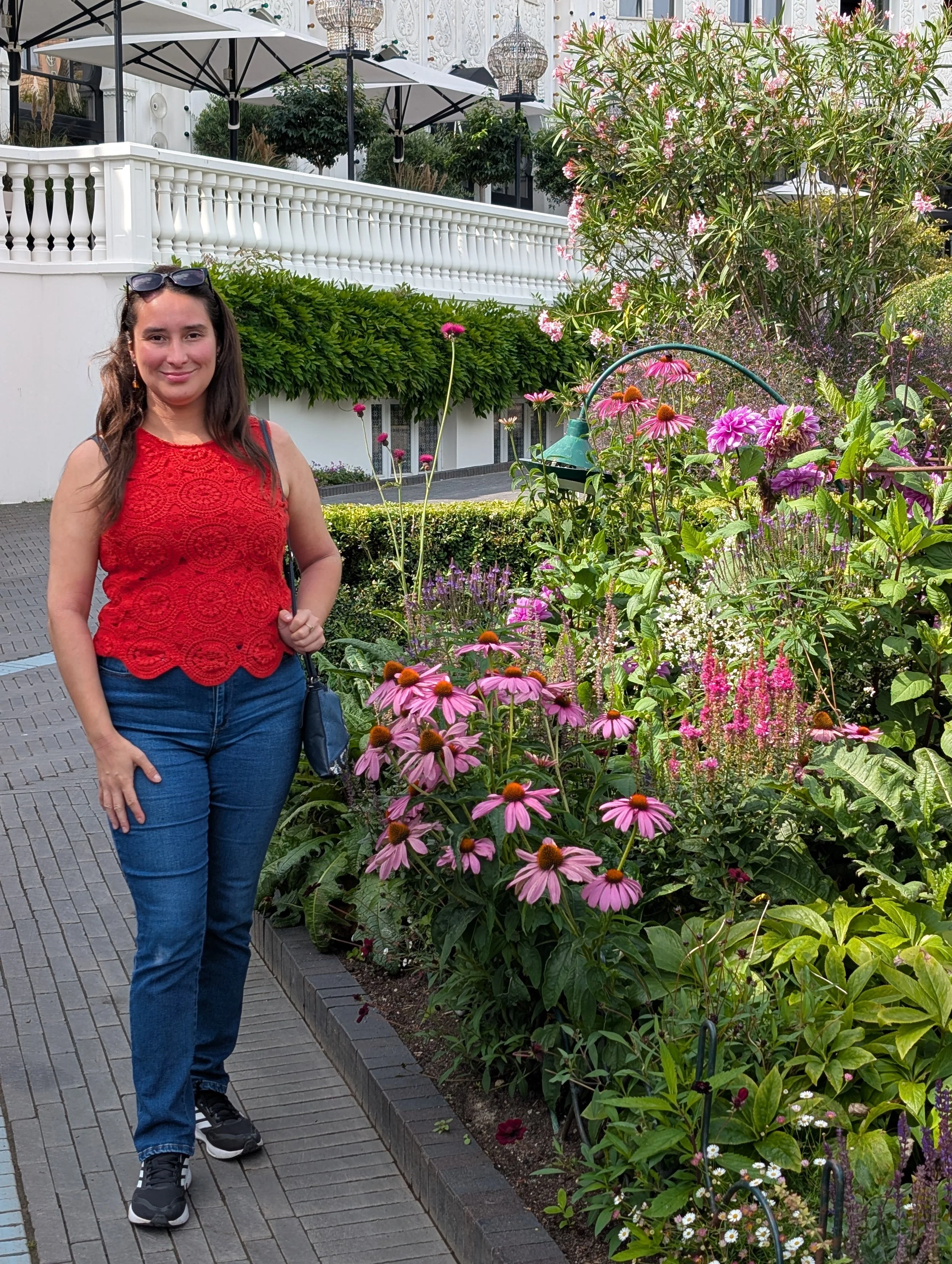 A woman with long brown hair wearing a red sleeveless top and blue jeans stands on a brick sidewalk surrounded by colorful flowers, with white building with black windows and white railings in the background.