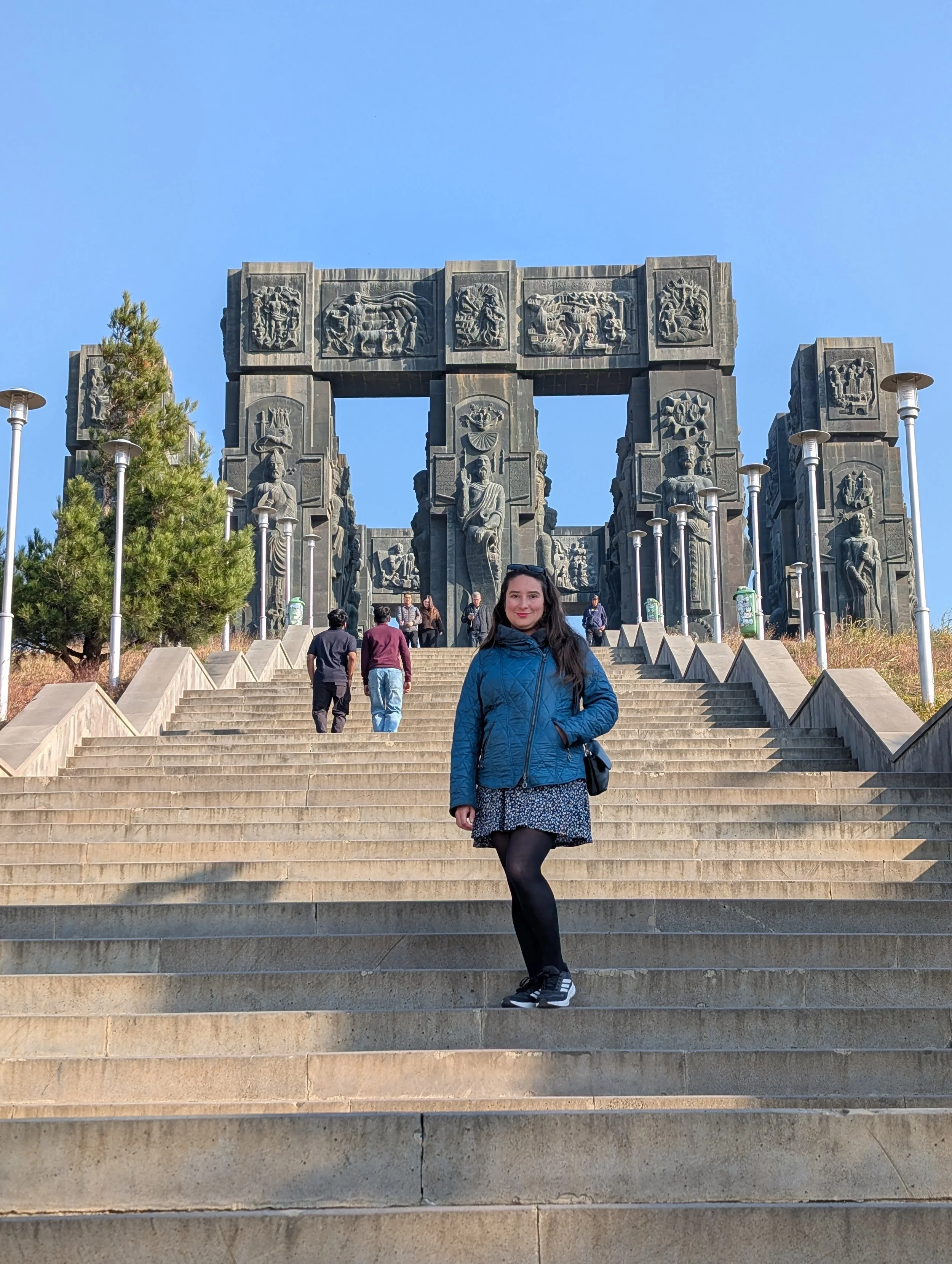 Woman in a blue jacket posing on wide stone stairs in front of a large monument with carved relief sculptures, under clear blue sky.