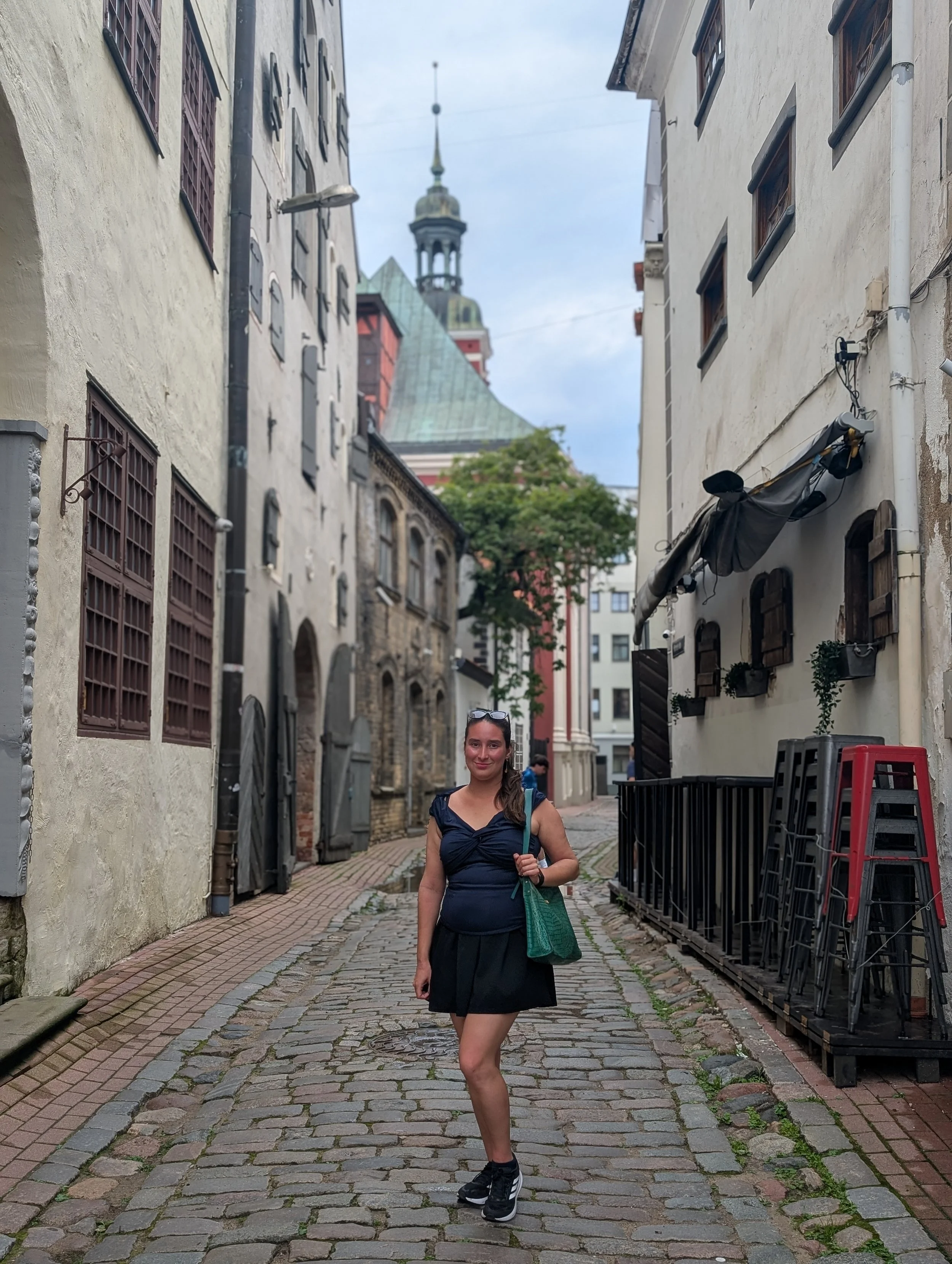 A woman with long brown hair, wearing a black dress and black sneakers, standing on a cobblestone street in a historic European city. She is holding a green bag and smiling at the camera. The street is lined with old buildings with barred windows and