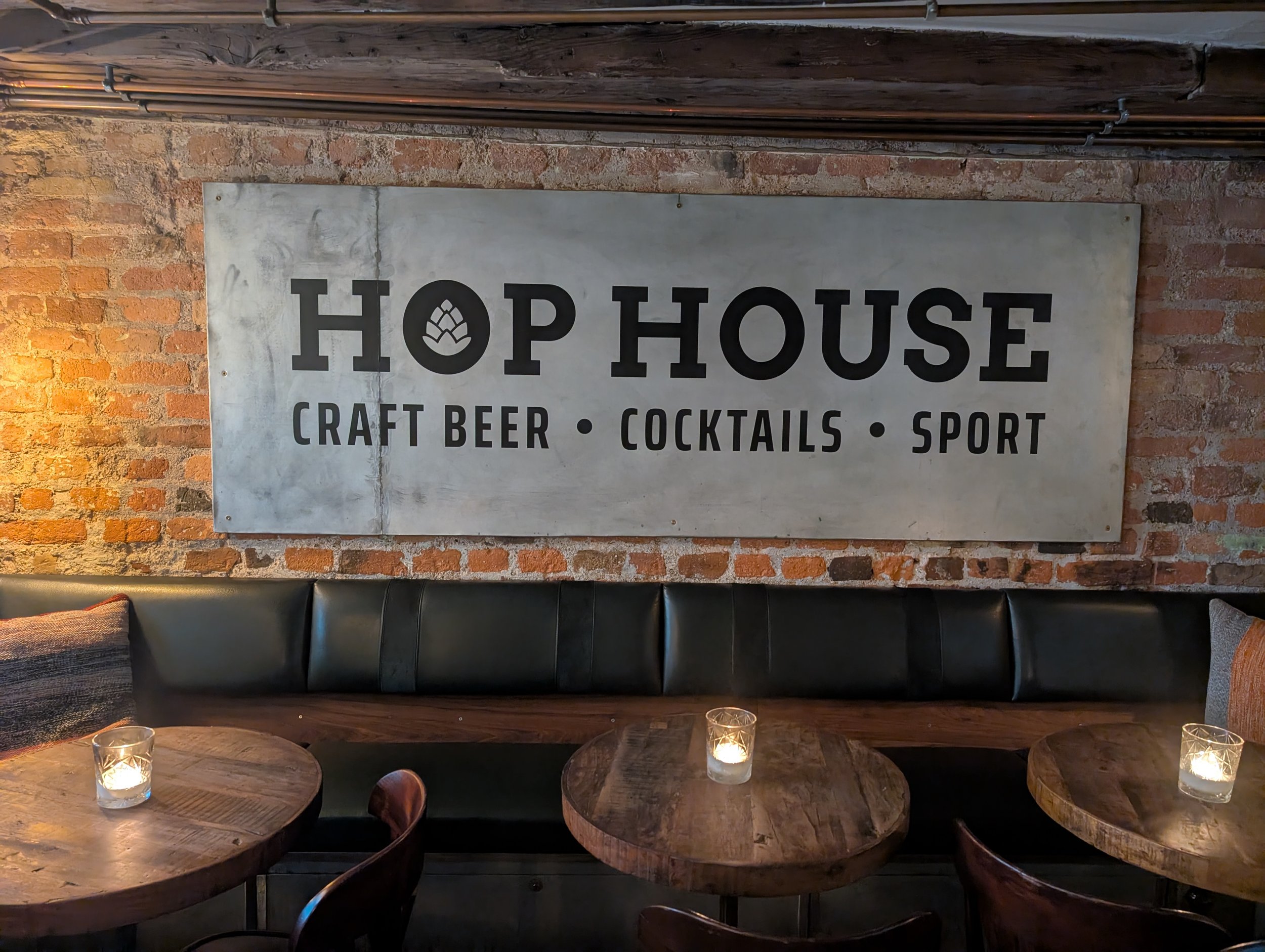 Interior of a restaurant or bar with a brick wall, a metal sign reading 'HOP HOUSE' with a hop cone logo, and a row of wooden tables with candles in glass holders.