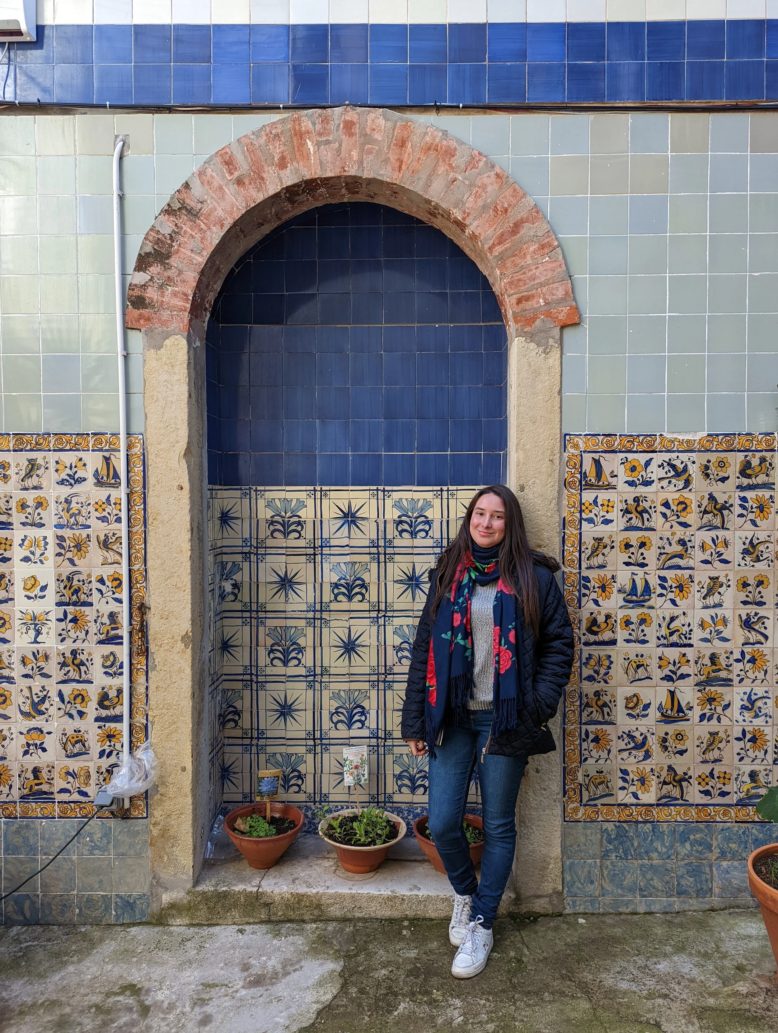 Young woman standing in front of a decorative tiled wall with potted plants, smiling at the camera.