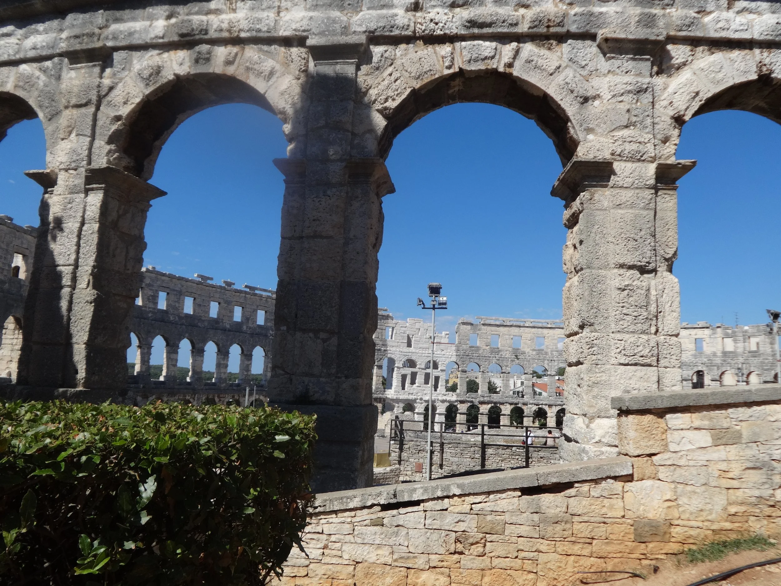 Ancient stone walls and arches of the Colosseum, with a clear blue sky and some modern structures and people visible in the background.