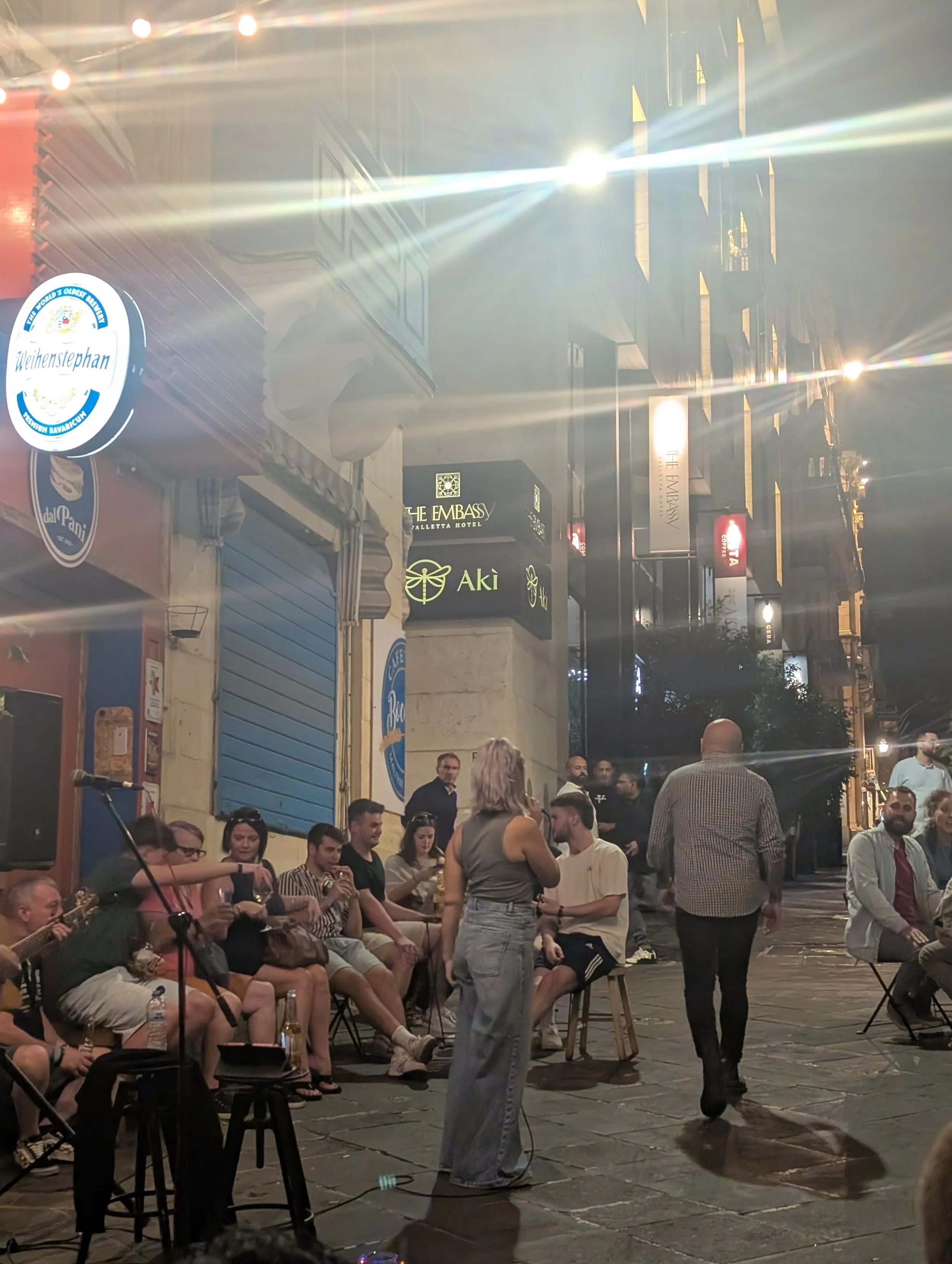 People sitting and standing on a street at night, some holding drinks, while a musician plays guitar. Streetlights and building signs are visible.