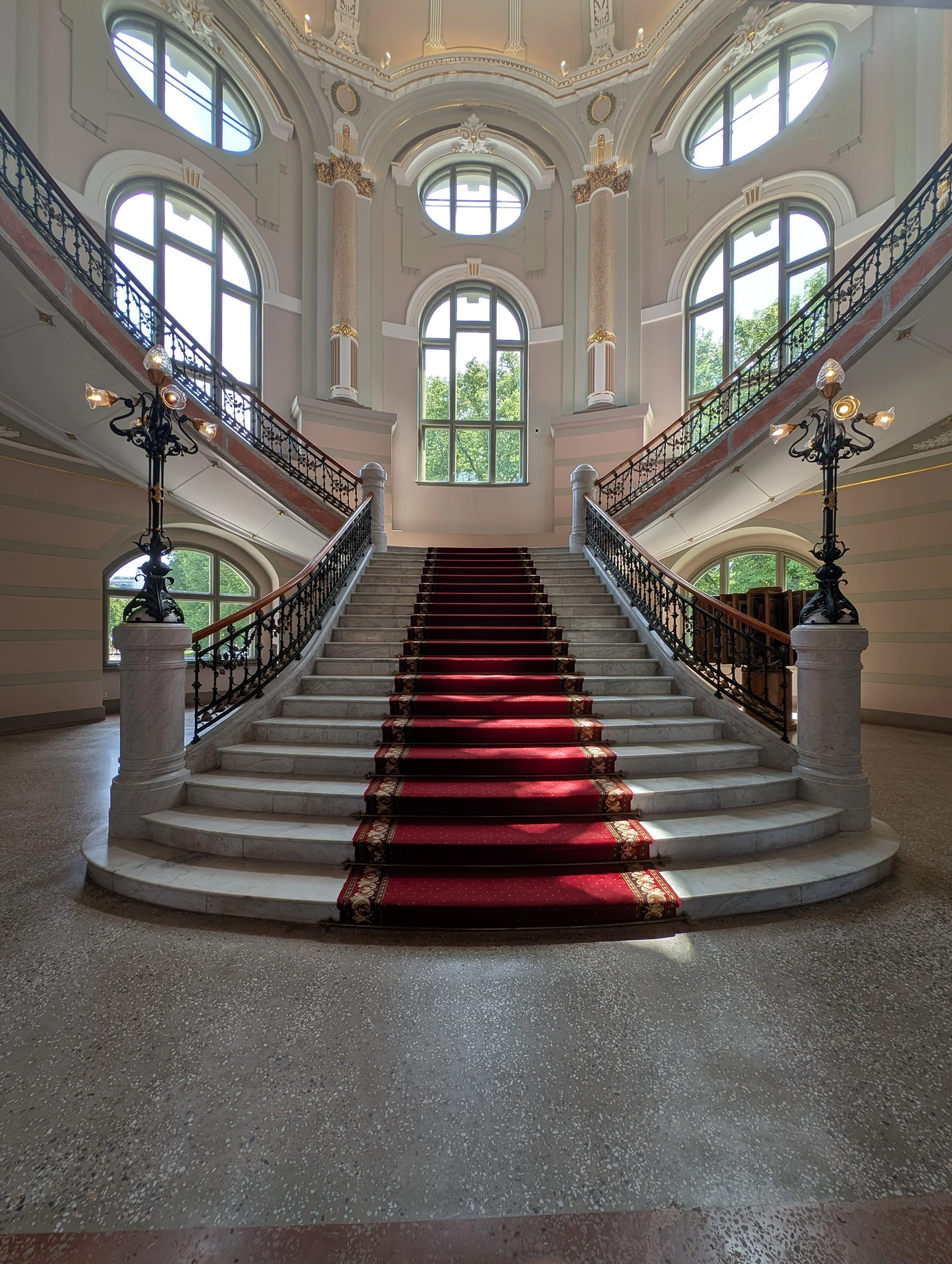 Grand staircase with marble steps, red carpet runner, ornate black wrought iron railing, and elegant ceiling with large windows and decorative gold accents, inside a historic building or palace.