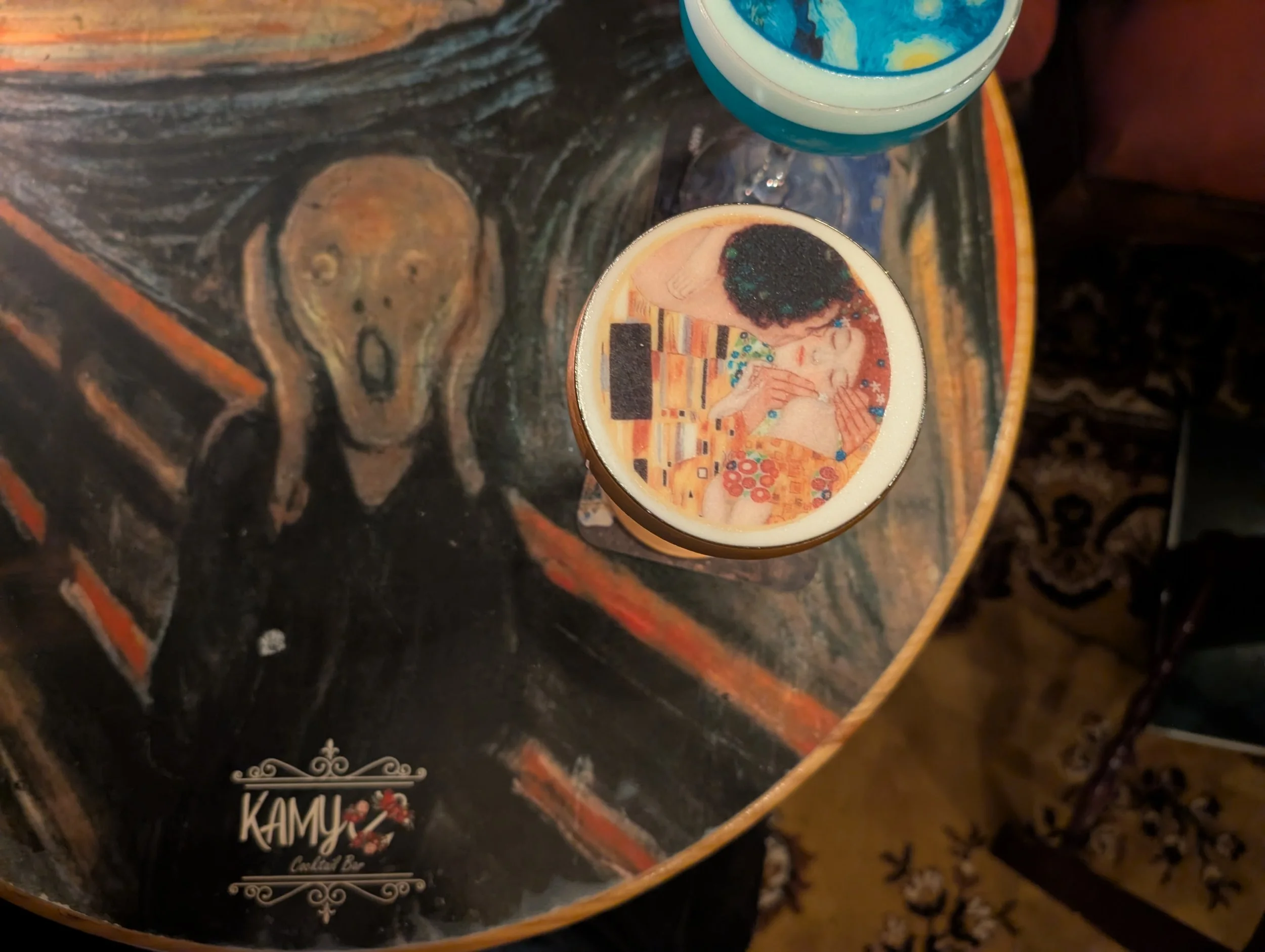 Two coffee cups placed on a dark, textured surface with a colorful, abstract painting underneath. One cup features an image from the painting on its surface.