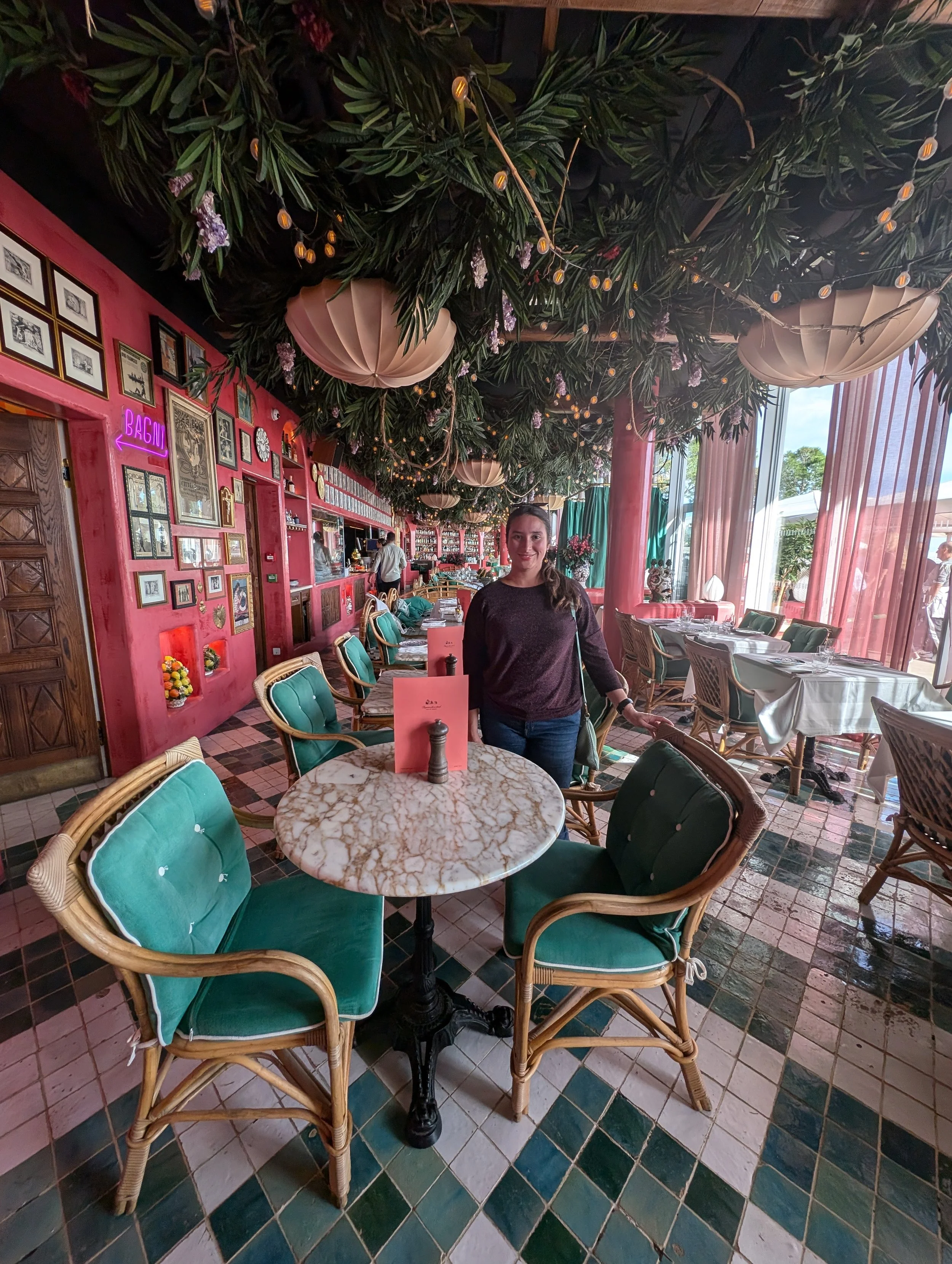 Interior of a restaurant with pink walls, framed pictures, and a woman standing near a white marble table with green cushioned chairs, large windows with curtains, and a decorated ceiling with greenery and hanging lights.