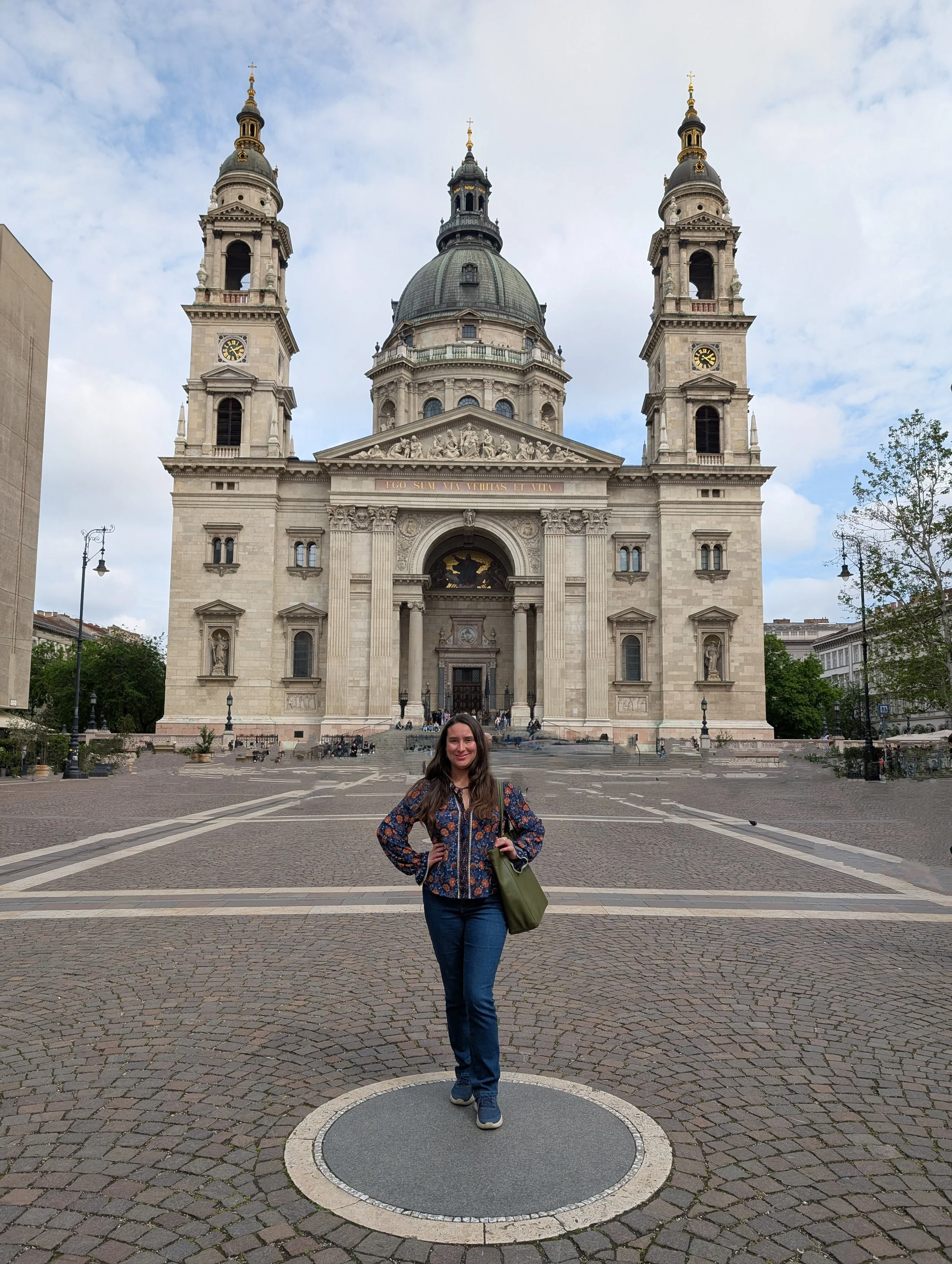 A woman in a patterned jacket and jeans standing with hands on hips in front of a large historic church with two bell towers, a dome, and an ornate pediment, on a cloudy day.