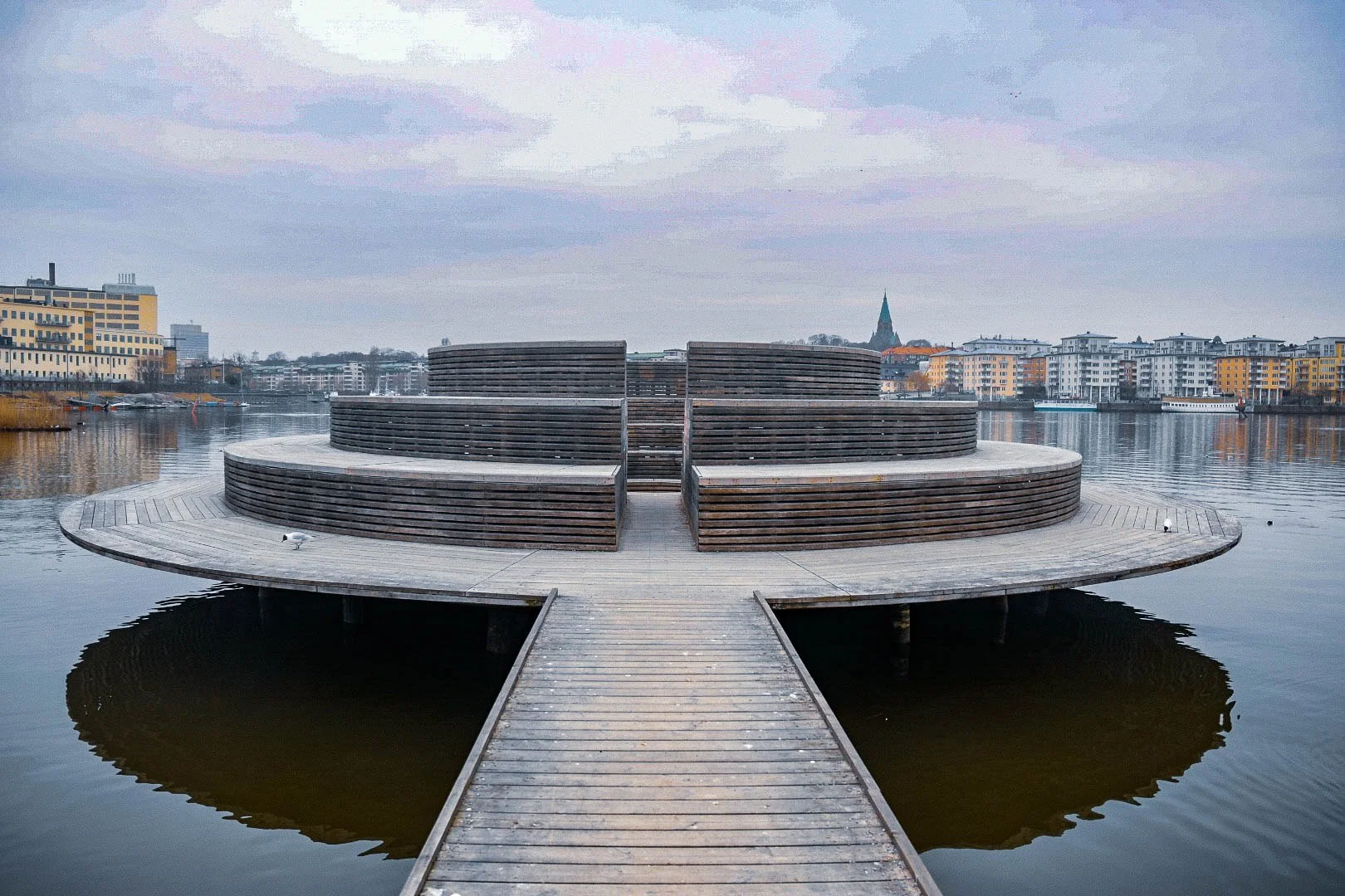 A modern wooden pier with a circular stage extending into a calm river, with city buildings and a church with a tall spire in the background.