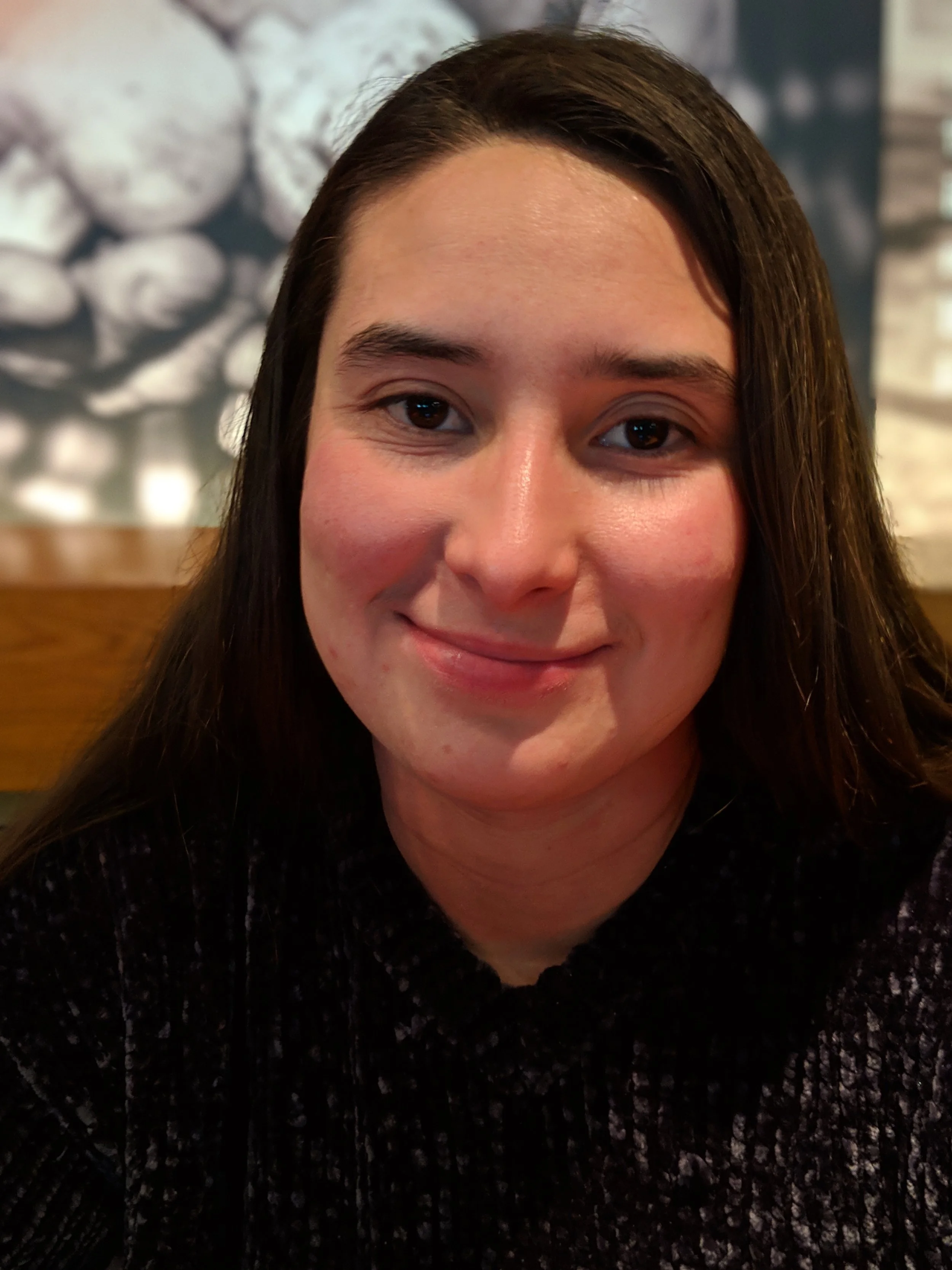 A woman with long dark hair smiling in a restaurant, with a blurred background of a wall decorated with what appears to be a decorative display of stones.