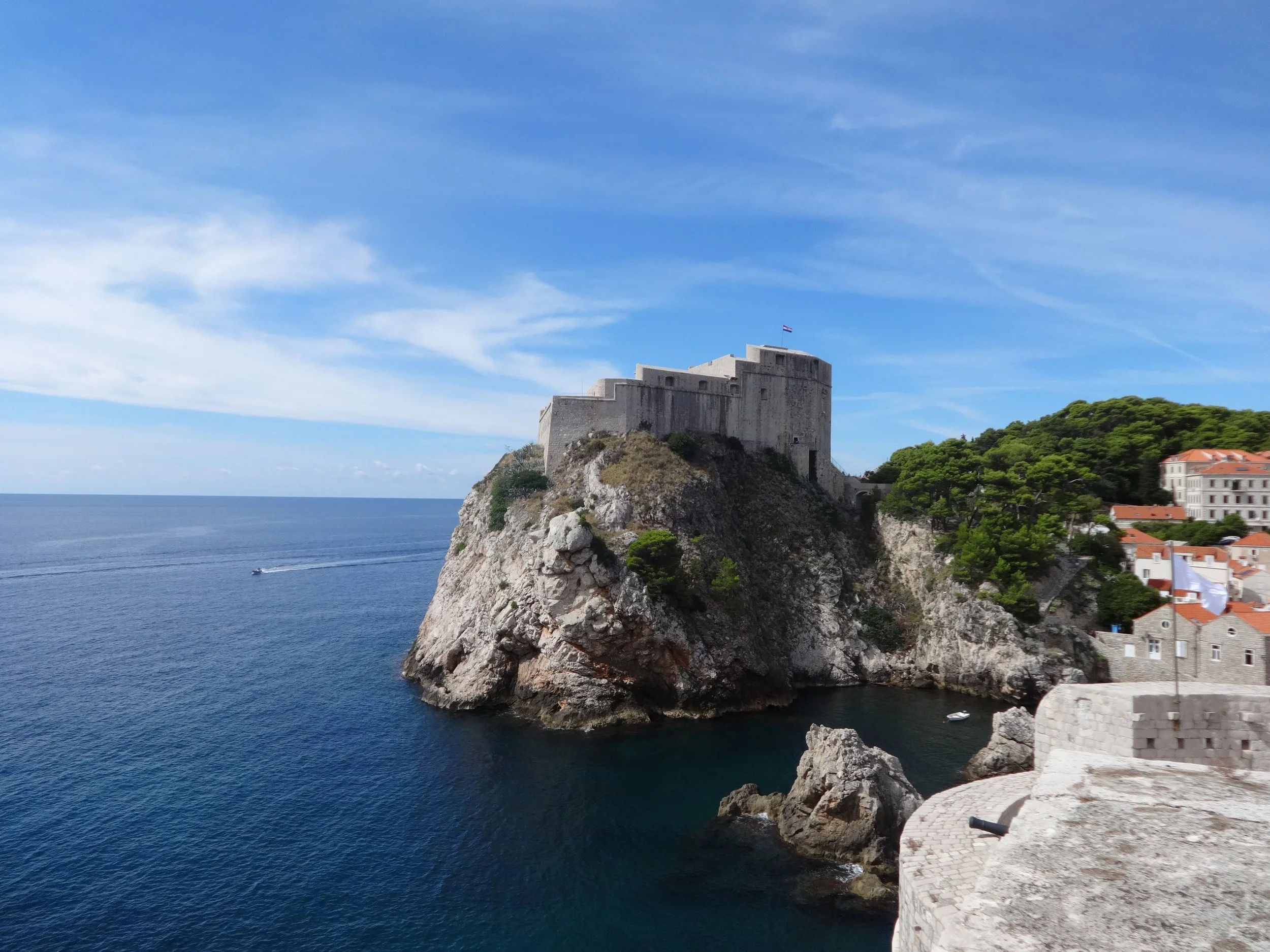 Castle on a rocky cliff overlooking the ocean with a cloudy blue sky in the background and trees on the right.