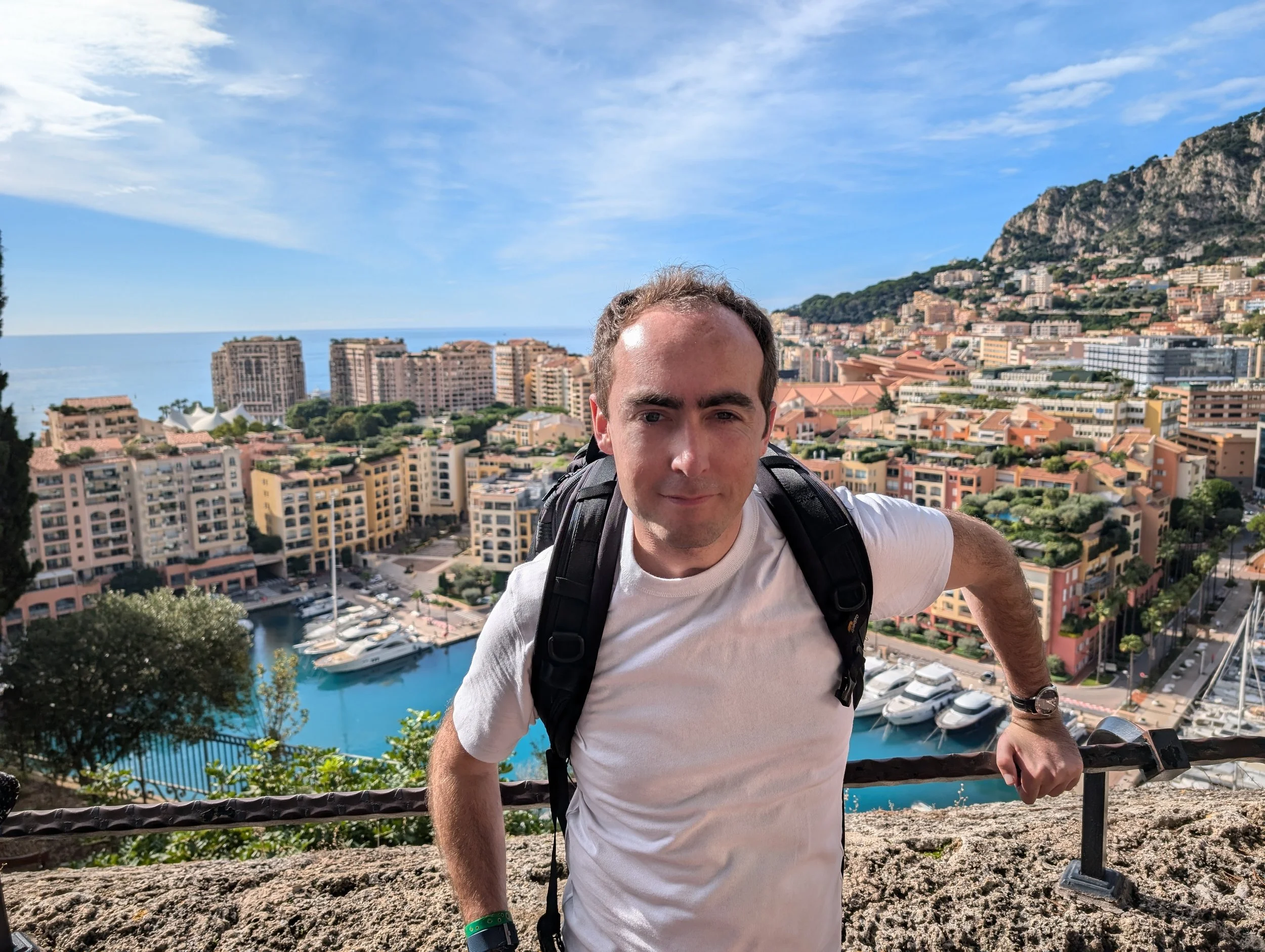 A young man wearing a white shirt and a black backpack, with outdoor gear on his wrist, standing on a scenic high vantage point overlooking a city with colorful buildings, a marina with yachts, and a mountainous landscape in the background under a pa