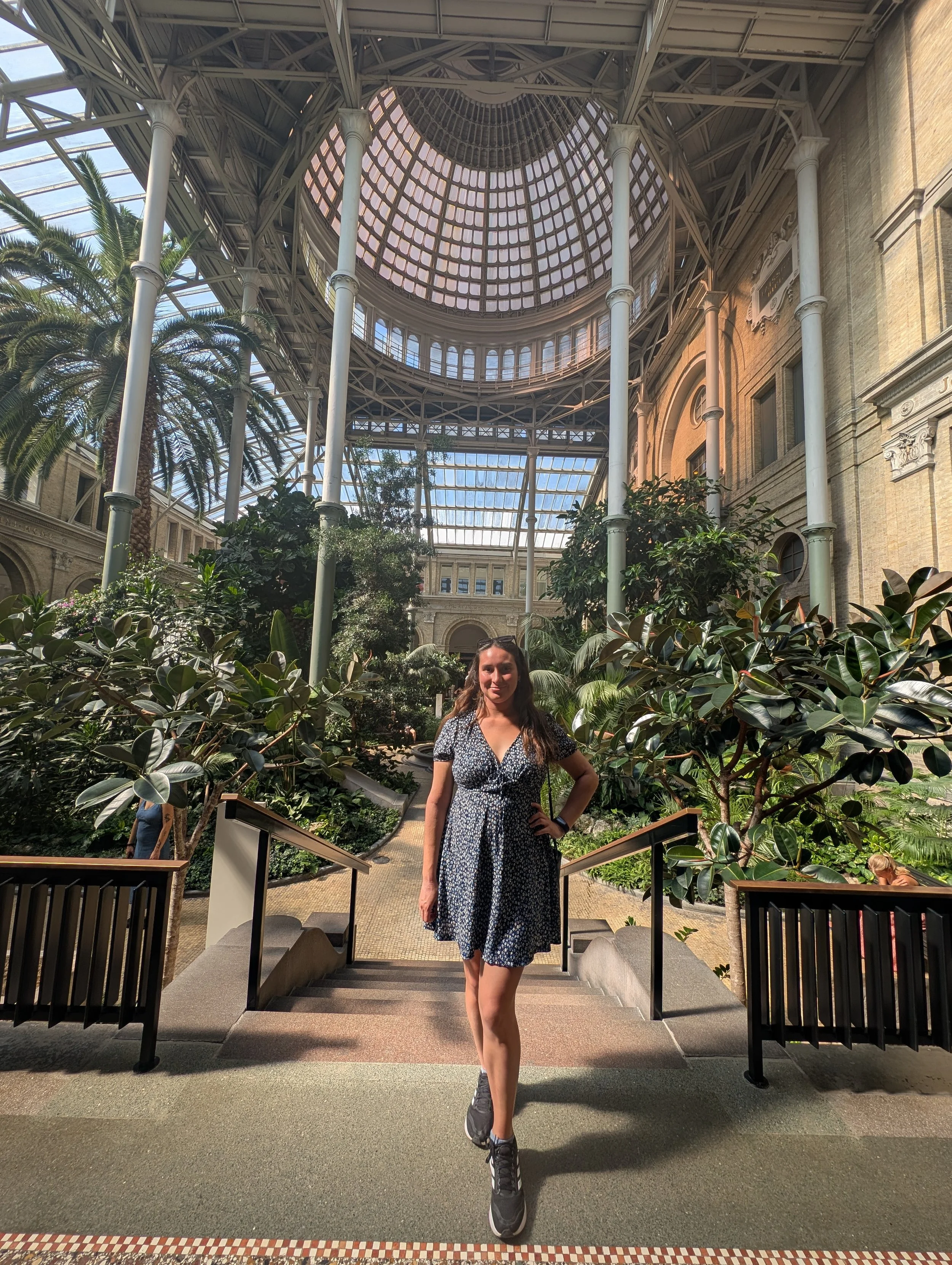 A woman standing on stairs inside a large, ornate glass-domed building filled with green plants. She is wearing a black and white floral dress and sneakers, smiling at the camera.