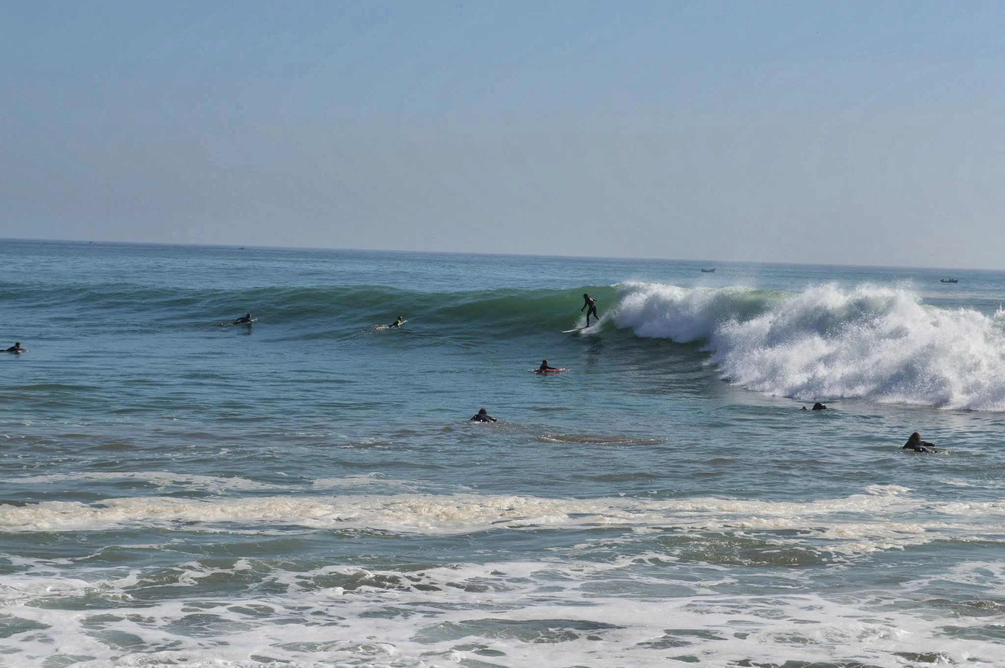 People surfing and swimming in the ocean near the shore with a clear sky and boats in the distance.