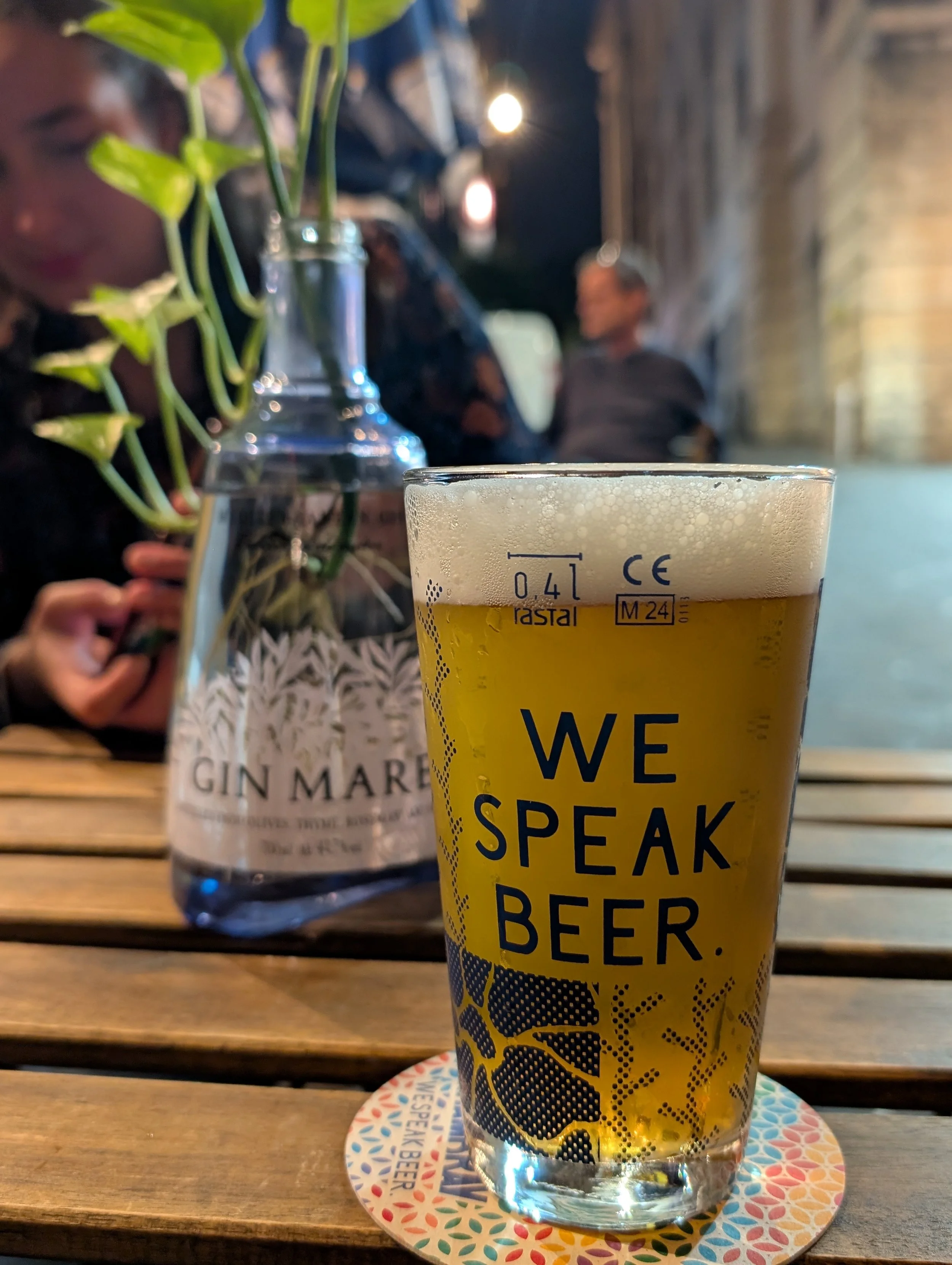 A pint of beer with a yellow label that reads "We Speak Beer," placed on a colorful patterned coaster on a wooden table. In the background, there are people socializing in a dimly lit setting with string lights and a brick wall.