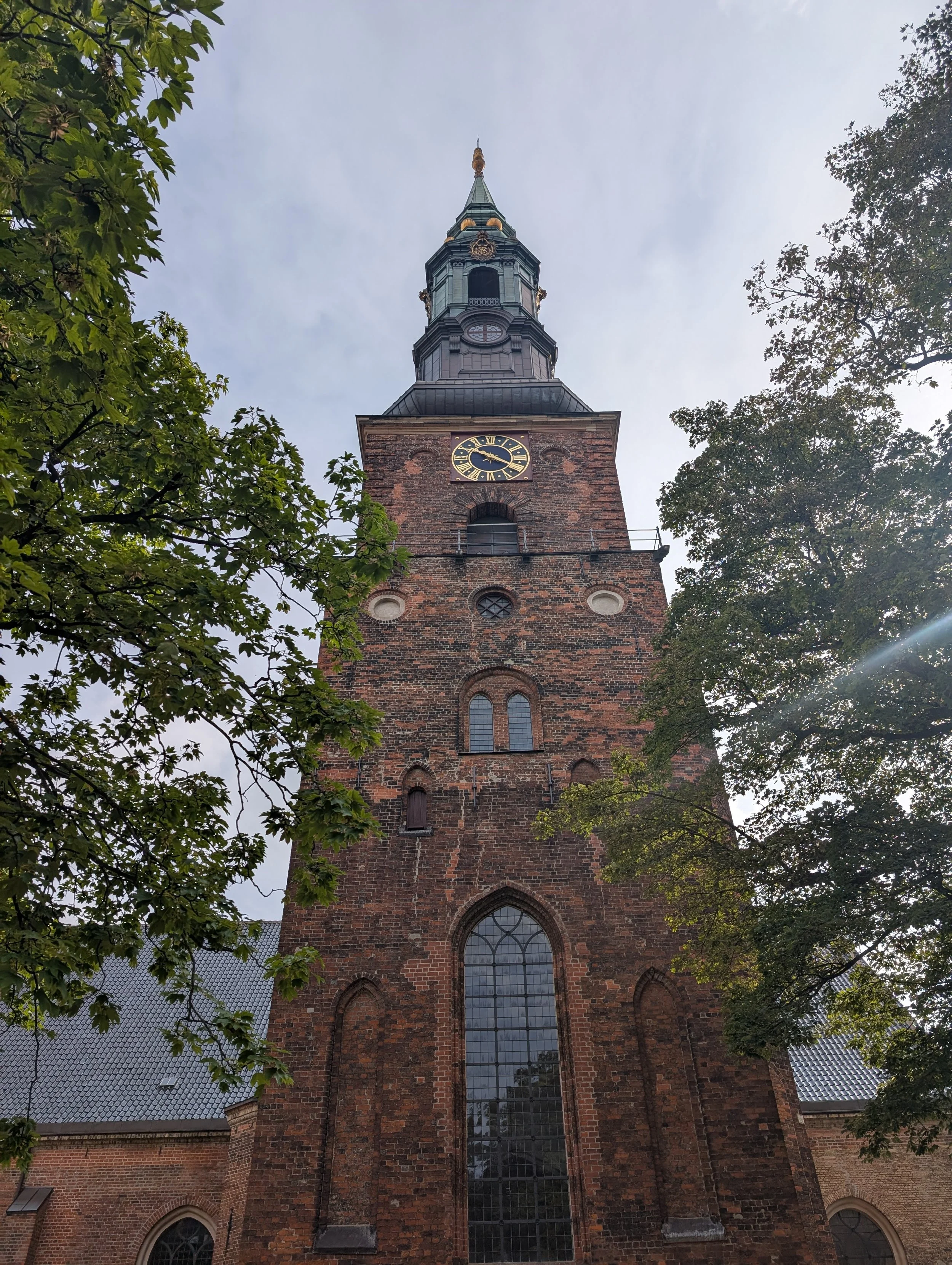A tall brick church tower with a clock and stained glass windows, surrounded by trees and an overcast sky.