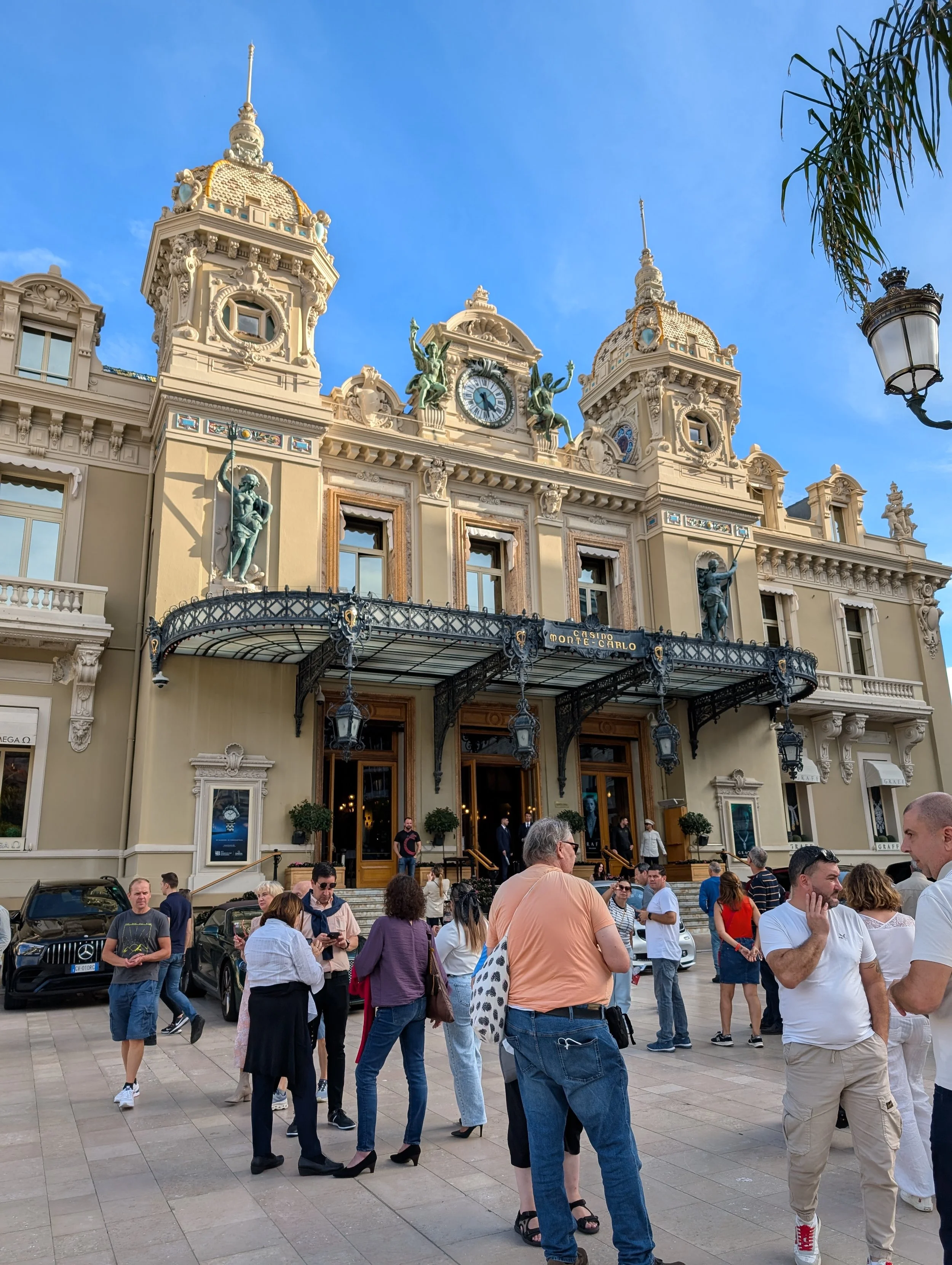 People gathering in front of a historic building with ornate architectural details, statues, and clock towers, under a blue sky with a few clouds.