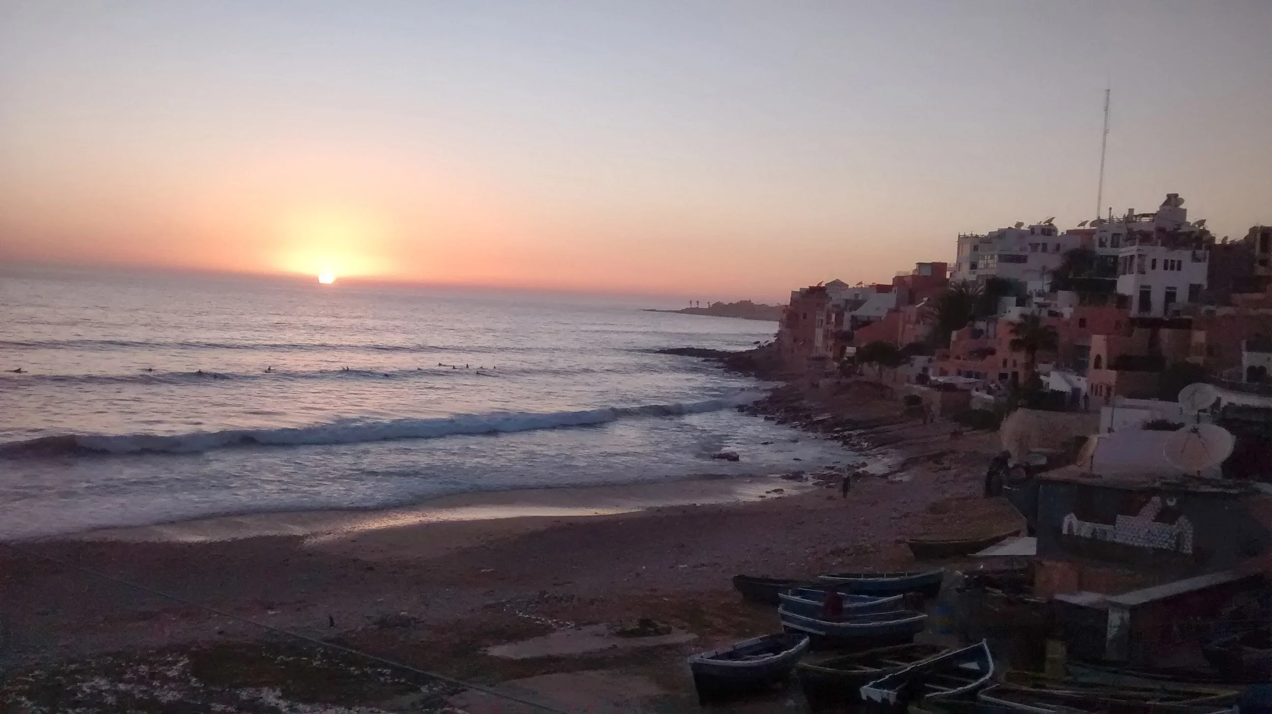 Sunset over the ocean with boats on the shore and houses on a hillside.