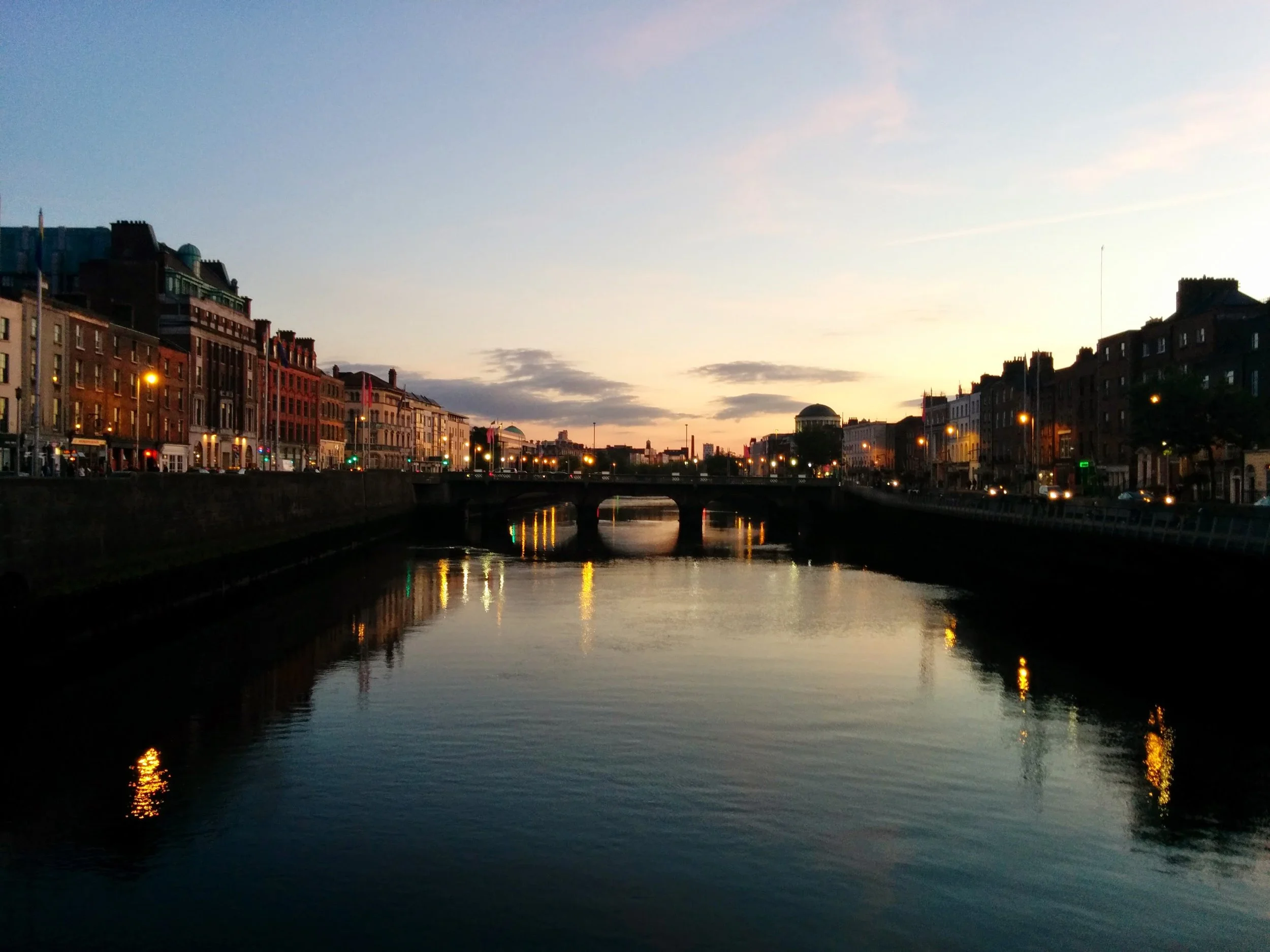 Sunset over a cityscape with buildings along a river, with reflections of lights on the water and a bridge crossing the river.