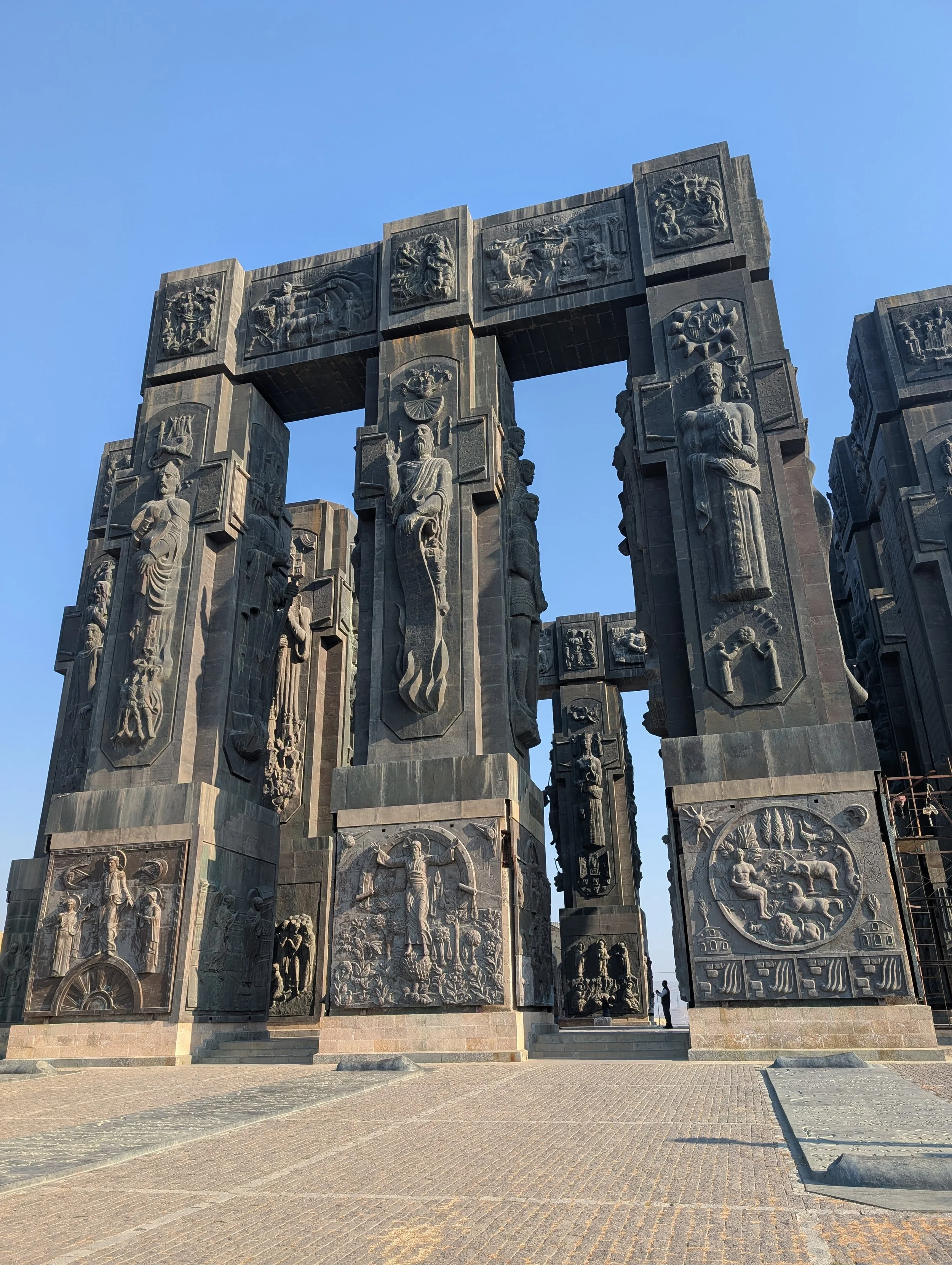 Large stone monument with intricate carved reliefs and figures, under a clear blue sky, in an open plaza.