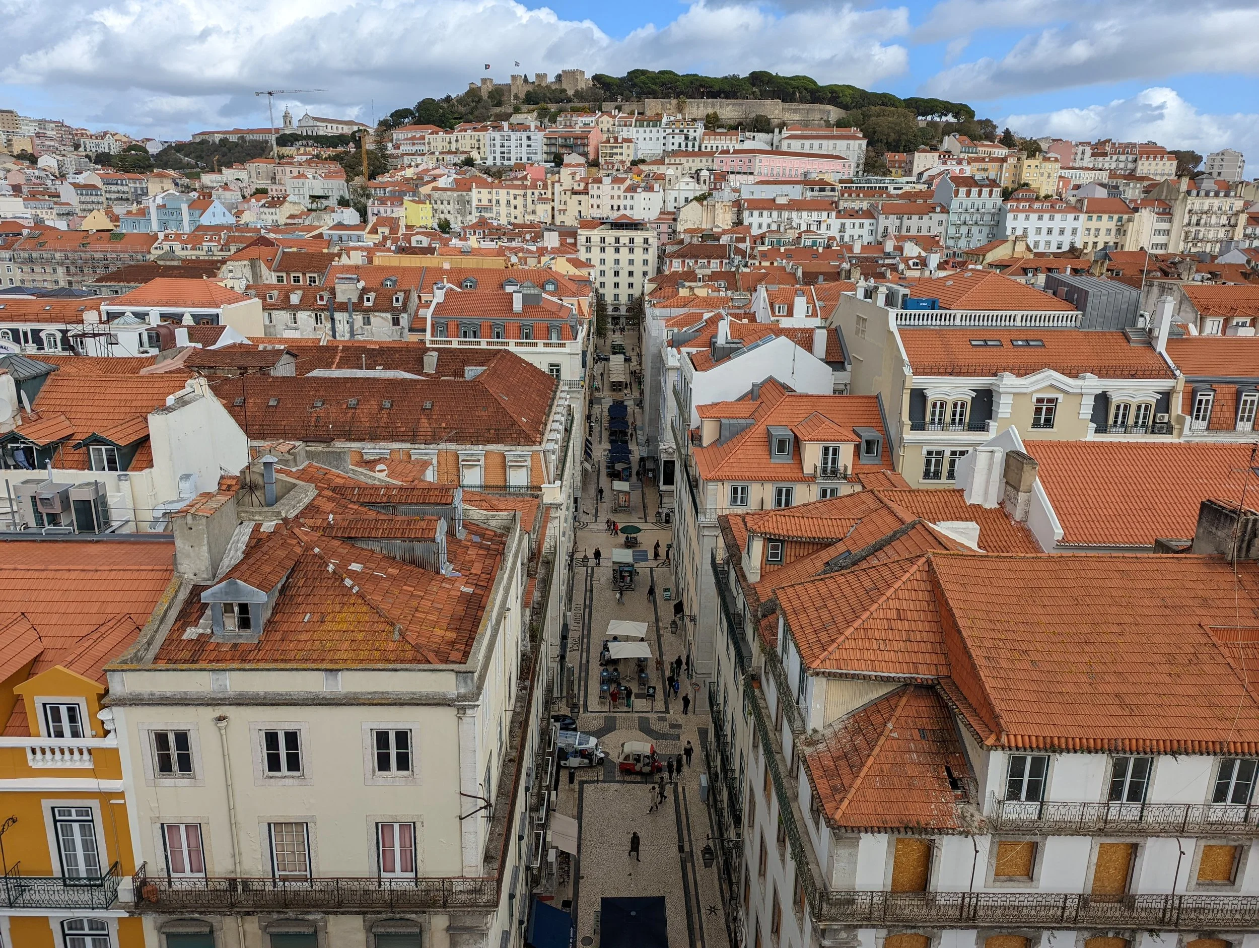 A cityscape of Lisbon, Portugal with orange-tiled roofs, narrow streets, and a hilltop castle in the background under a partly cloudy sky.