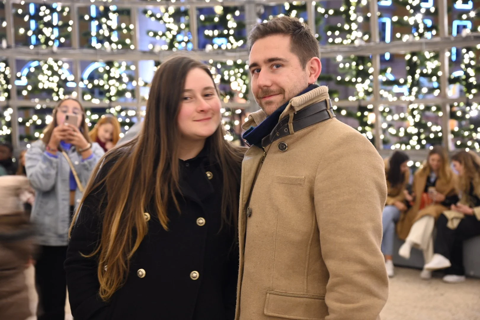 A couple posing in front of festive Christmas lights and decorated trees, with onlookers in the background.