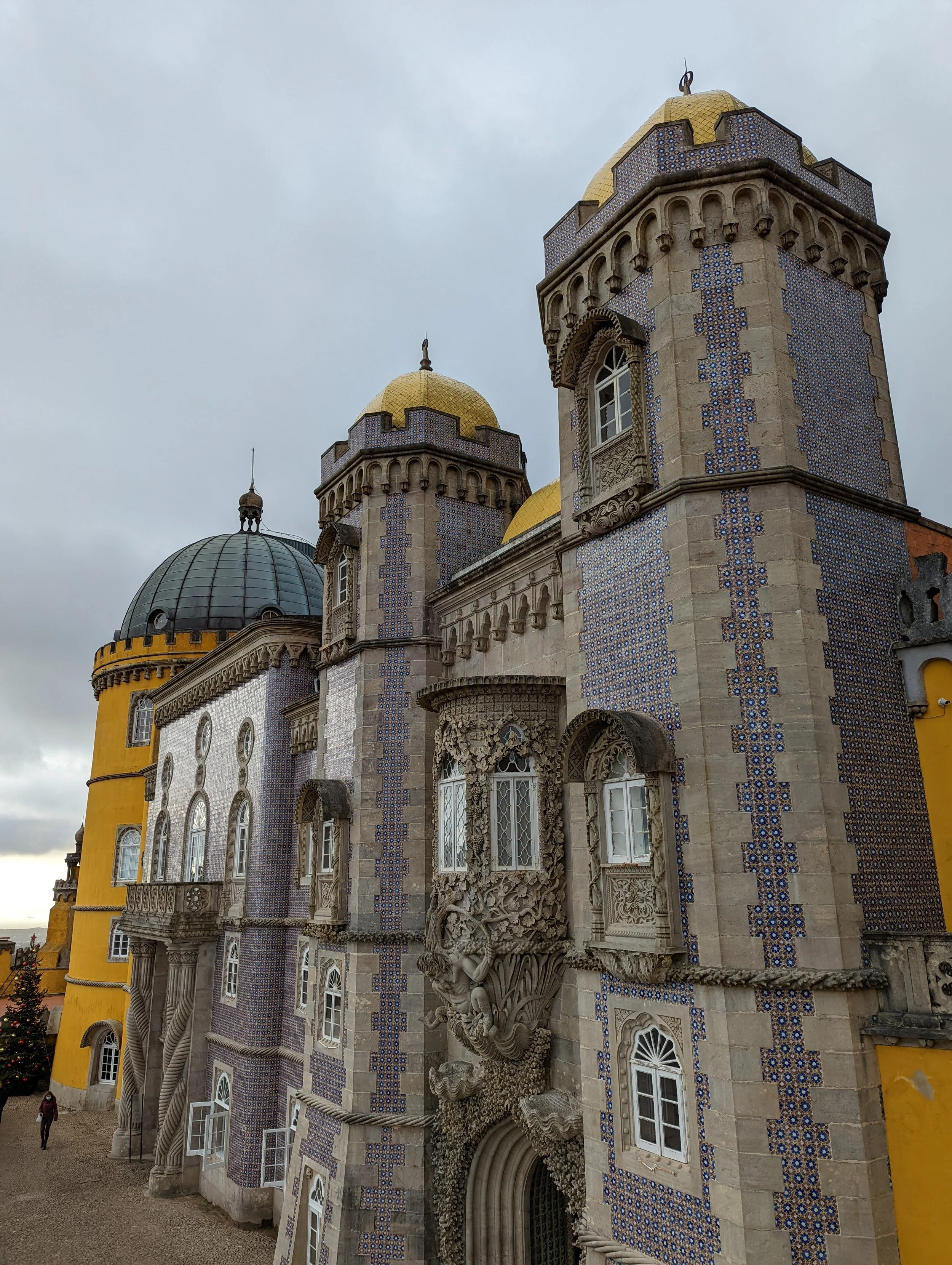 View of a castle-like building with ornate stonework, decorative blue and white tiles, and golden domed towers against a cloudy sky.