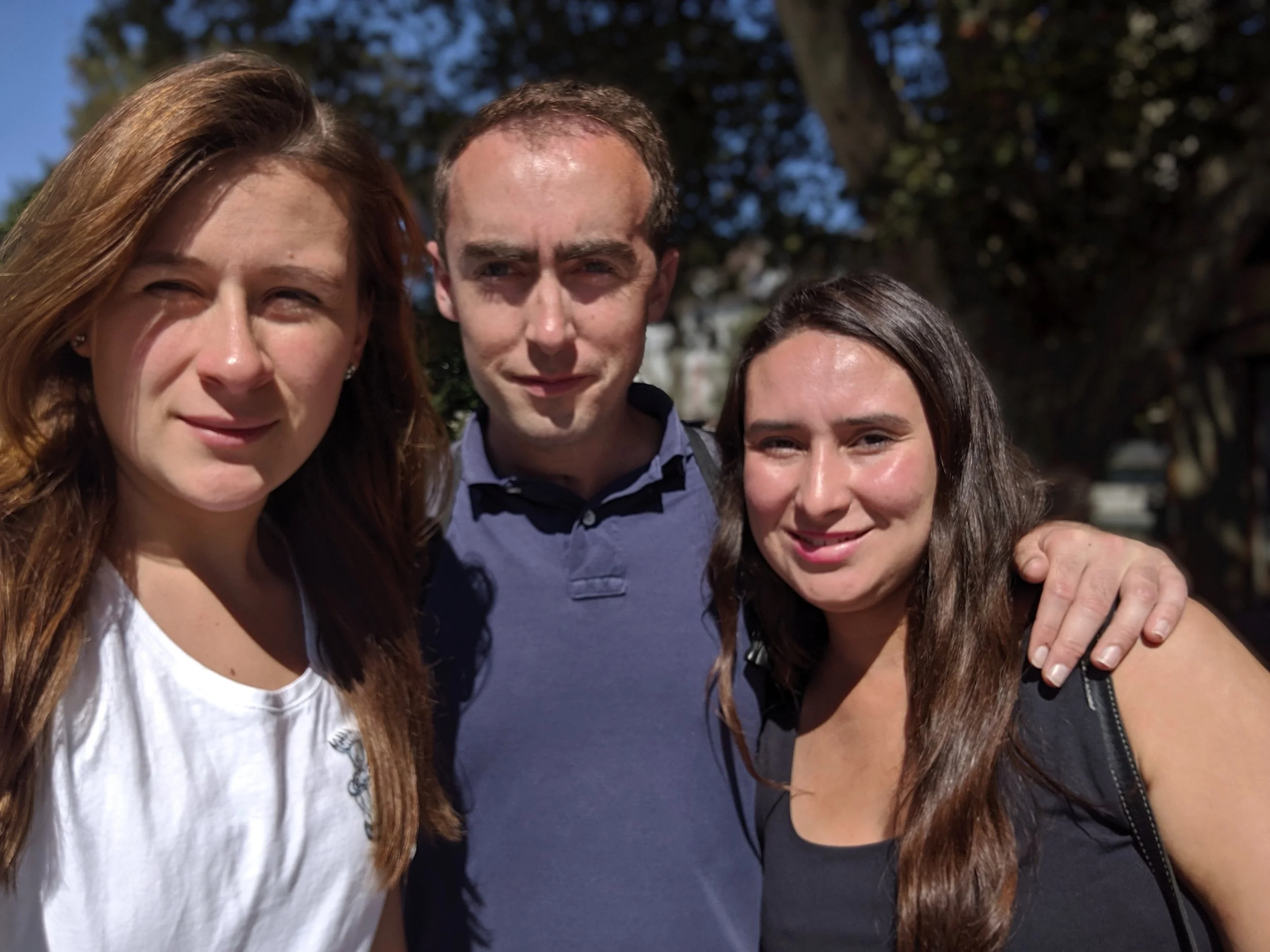 Three people, two women and one man, standing outdoors, smiling, with trees and a blue sky in the background.