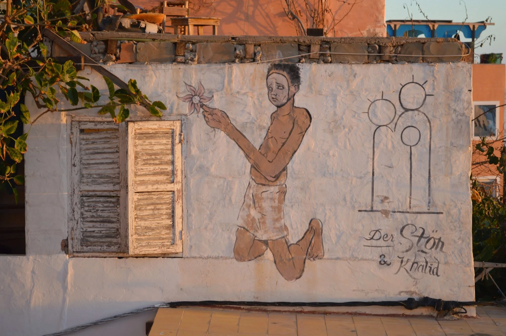 A mural of a boy holding a flower painted on a white wall is shown. The mural also includes simple black line drawings of three figures and some text in German reading "Der Sto" and "& Khalid." There is an old, weathered window shutter and some green