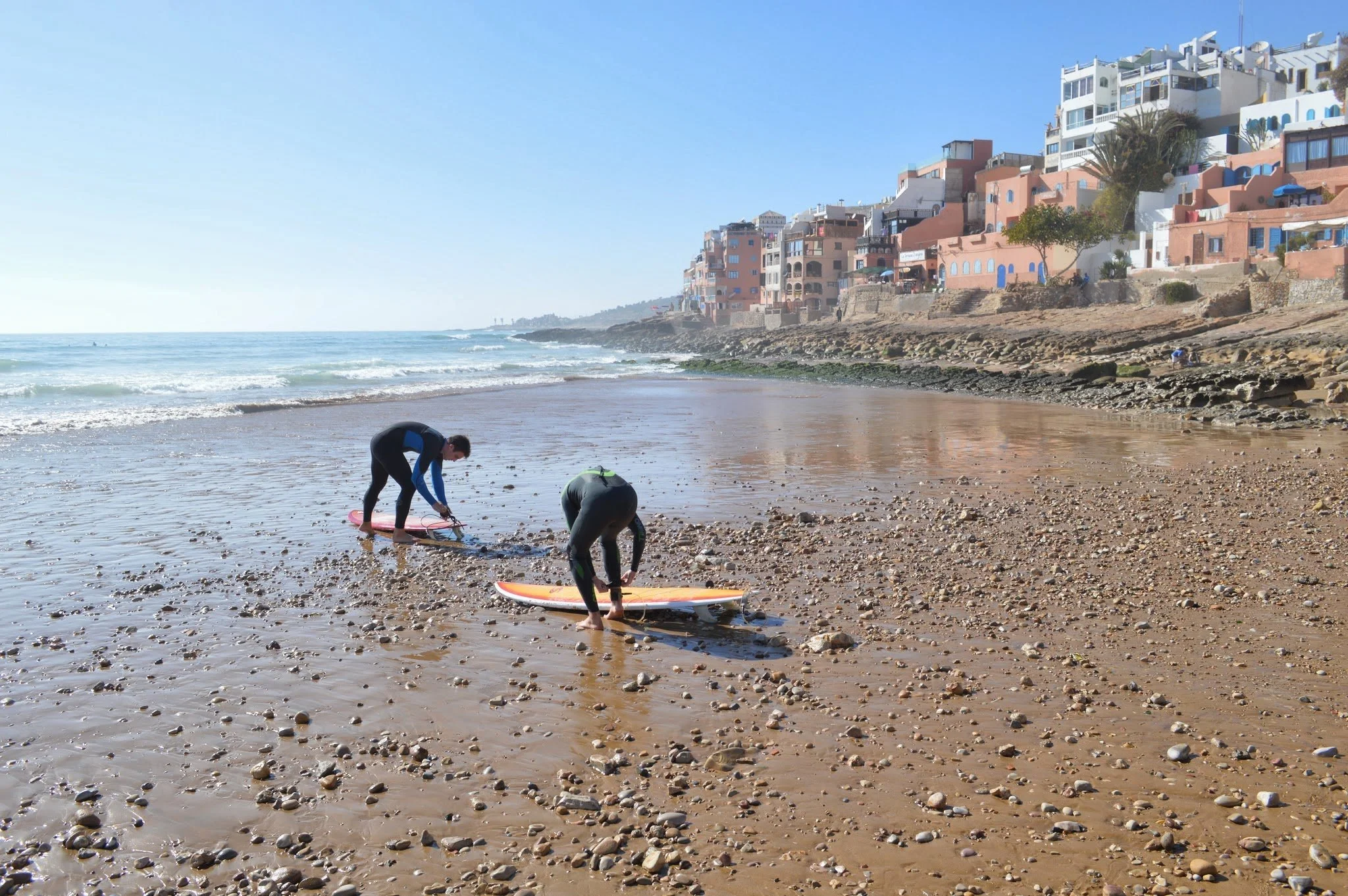 Two people in wetsuits preparing surfboards on a rocky beach near a coastal town with colorful buildings, under a clear blue sky.