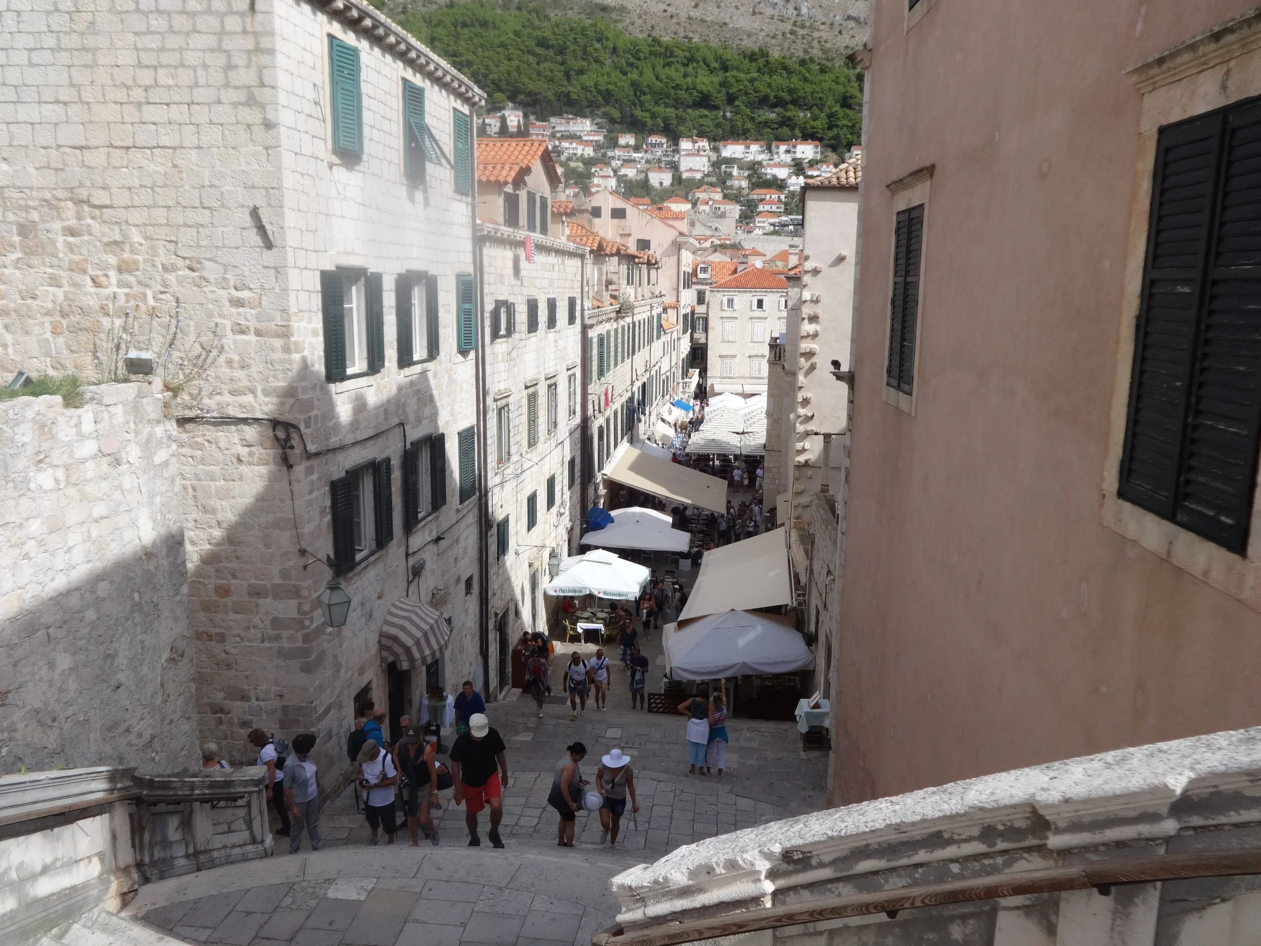 A narrow European street with stairs and outdoor cafes, crowded with tourists, surrounded by old stone buildings with shutters, against a backdrop of green hills with houses.