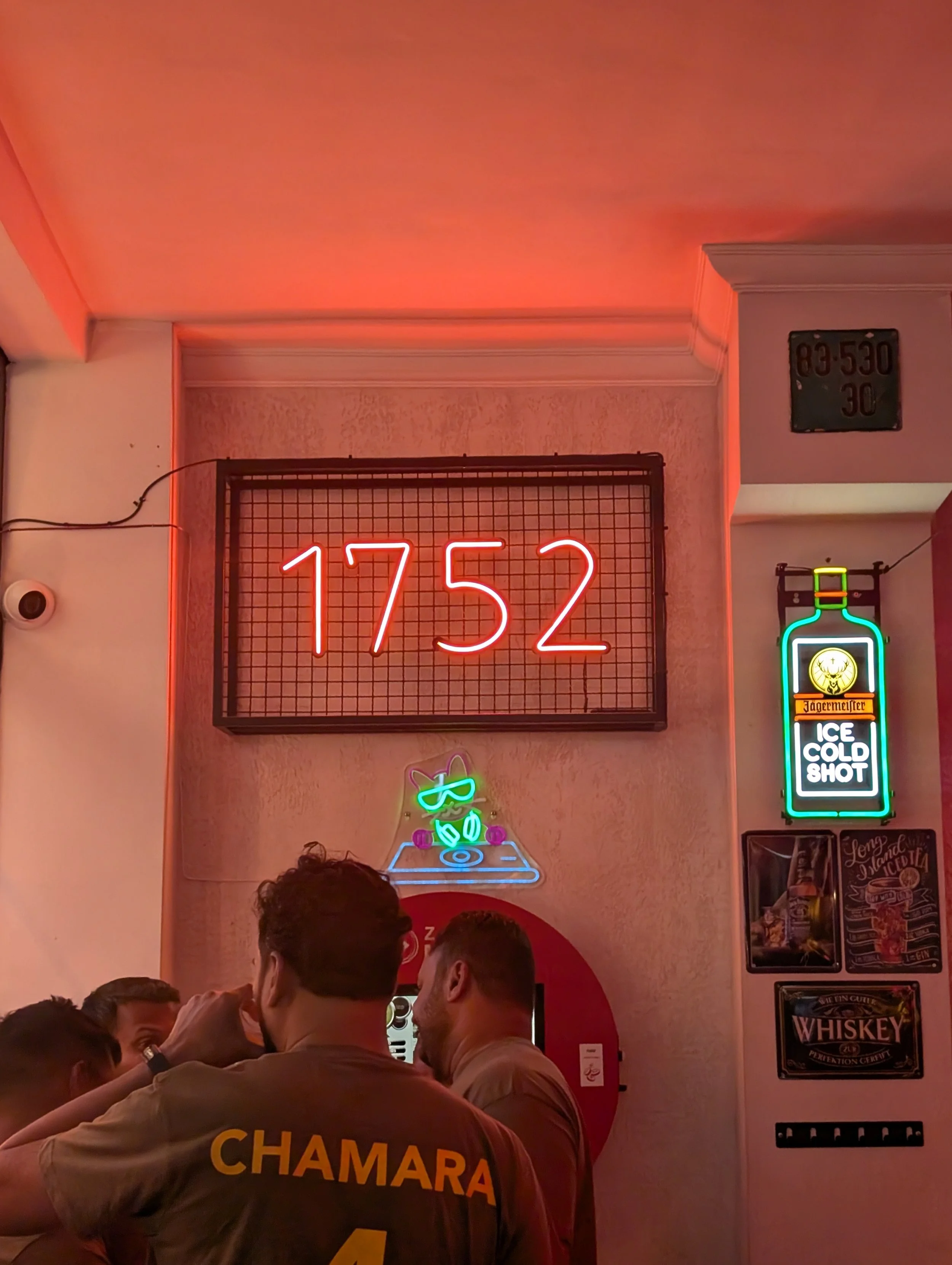 People standing in a line inside a restaurant with neon signs on the wall, including a large red number '1752' and other signs advertising drinks and food.