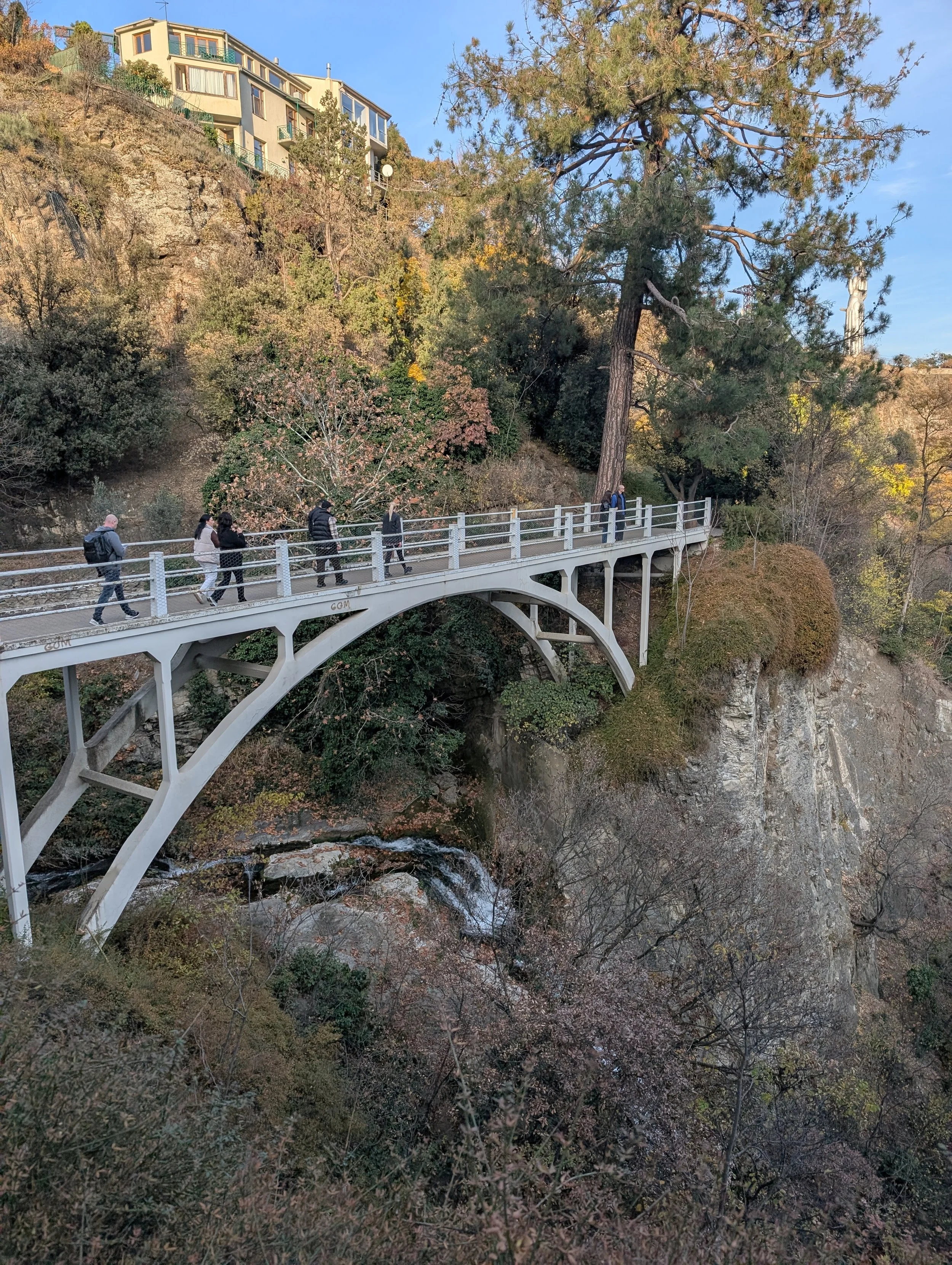 A group of people walking across a bridge over a rocky creek in a forested area with trees, bushes, and a hillside with houses.