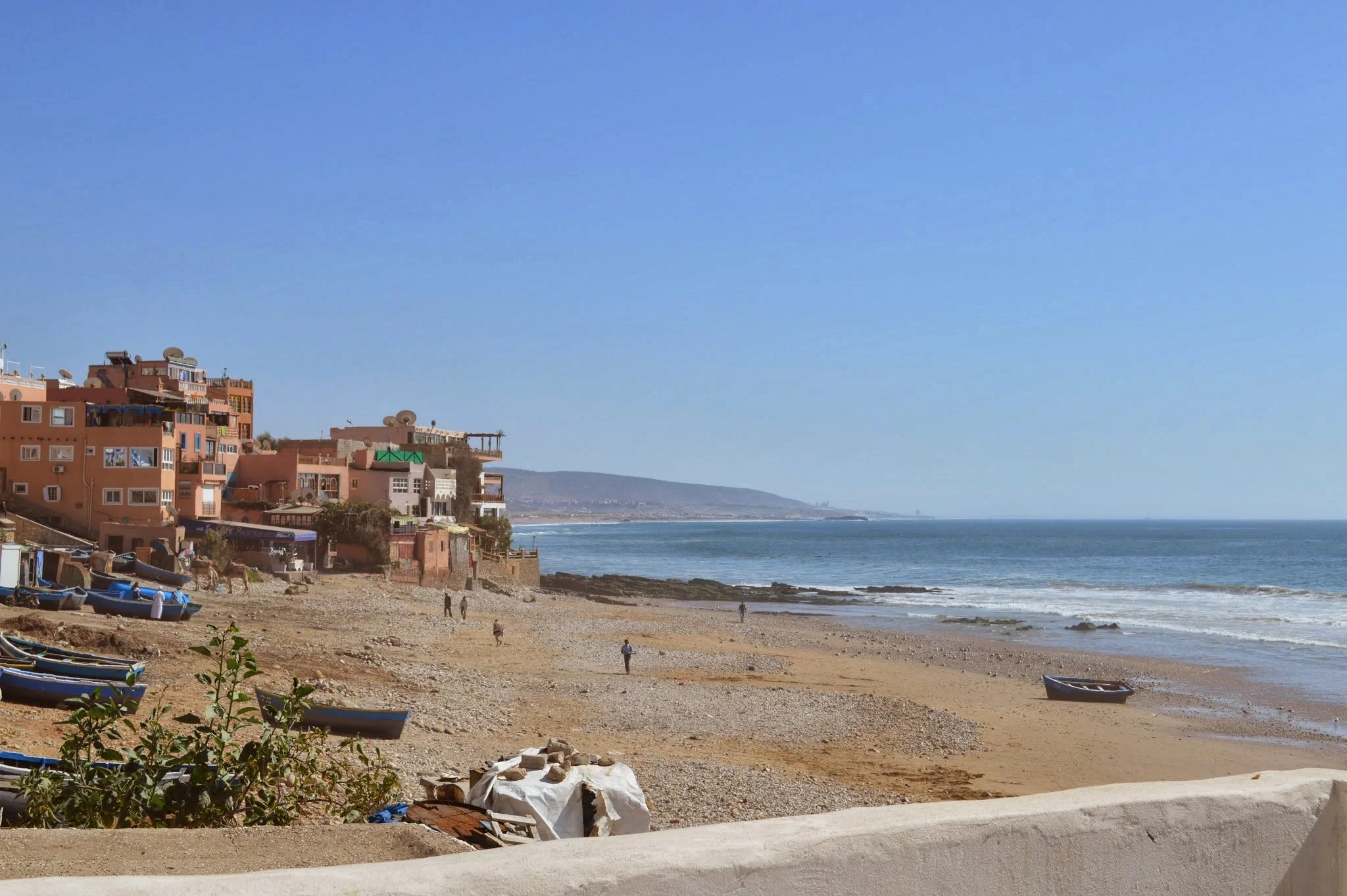 Beachside view with colorful buildings, boats on the sand, and the ocean in the background under a clear blue sky.