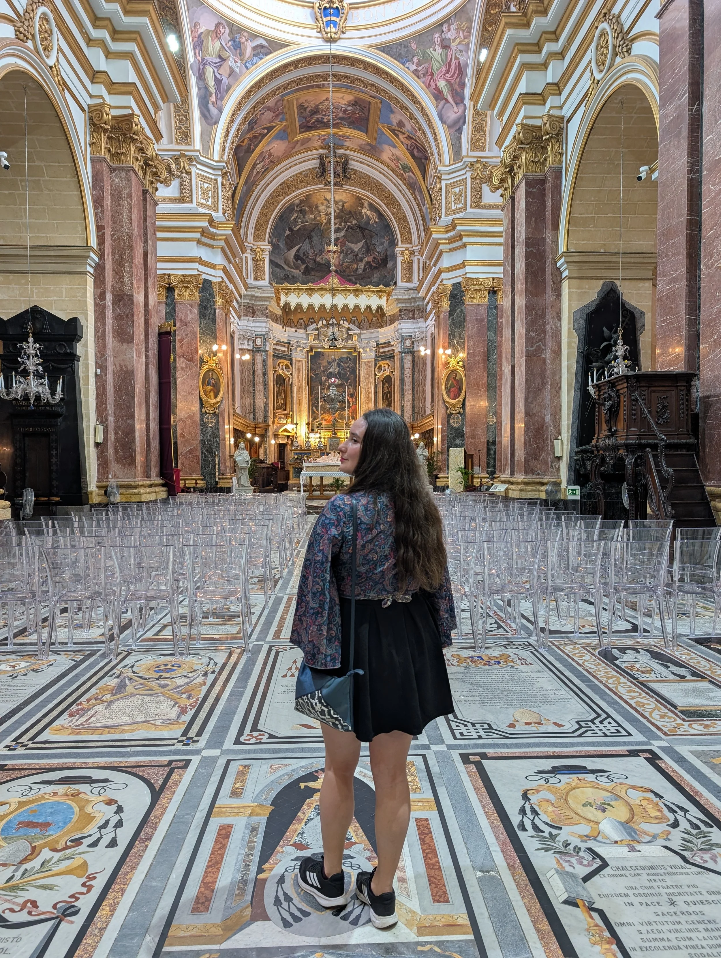 Inside a grand, ornate church or cathedral with elaborate gold and marble decorations, colorful painted ceilings, statues, and religious artwork. A woman with long, dark hair, wearing a patterned blouse, black skirt, and sneakers, stands among rows o