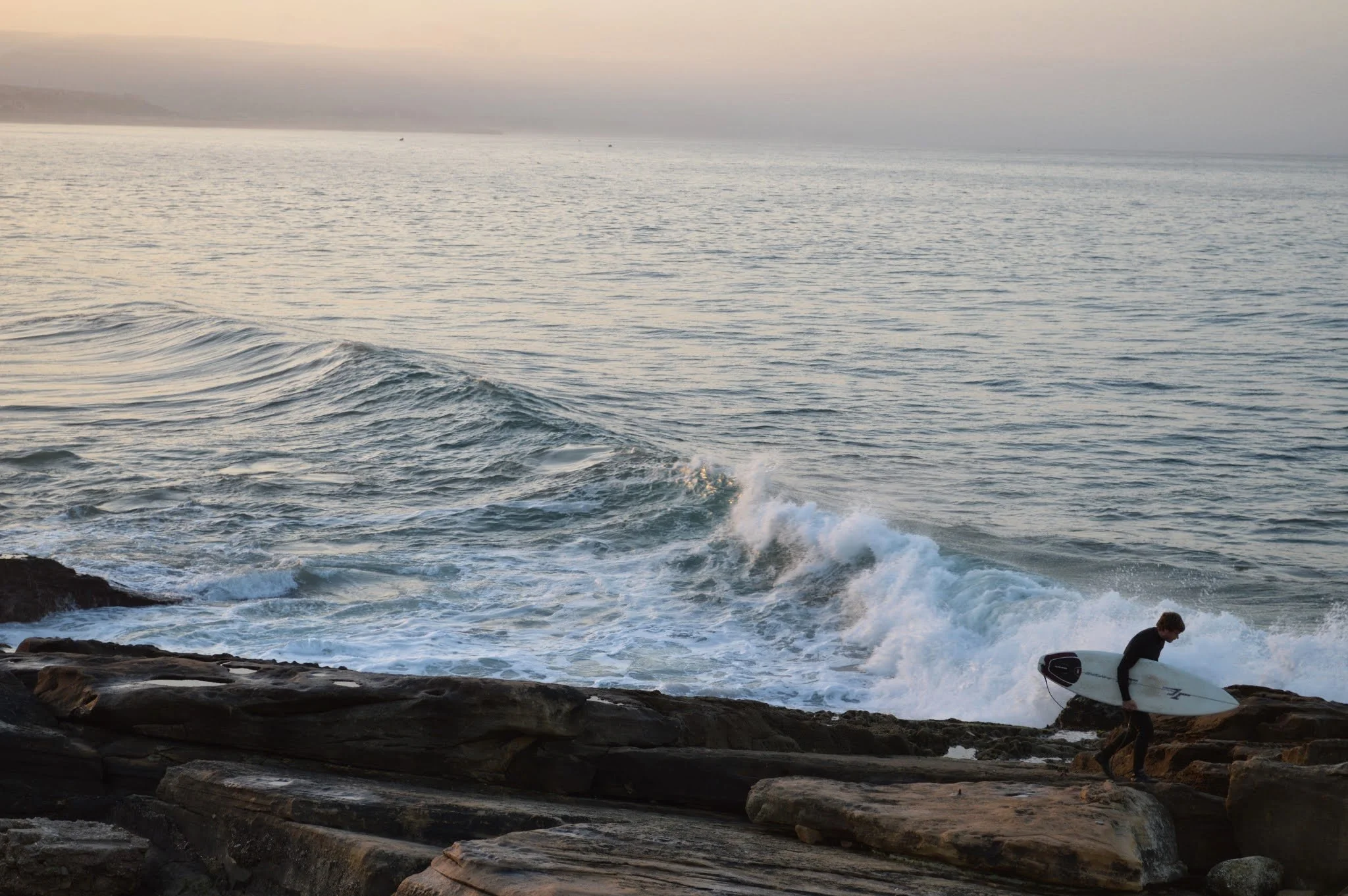 A person setting up a surfboard on rocky shore with the ocean and waves in the background, during dusk or dawn.