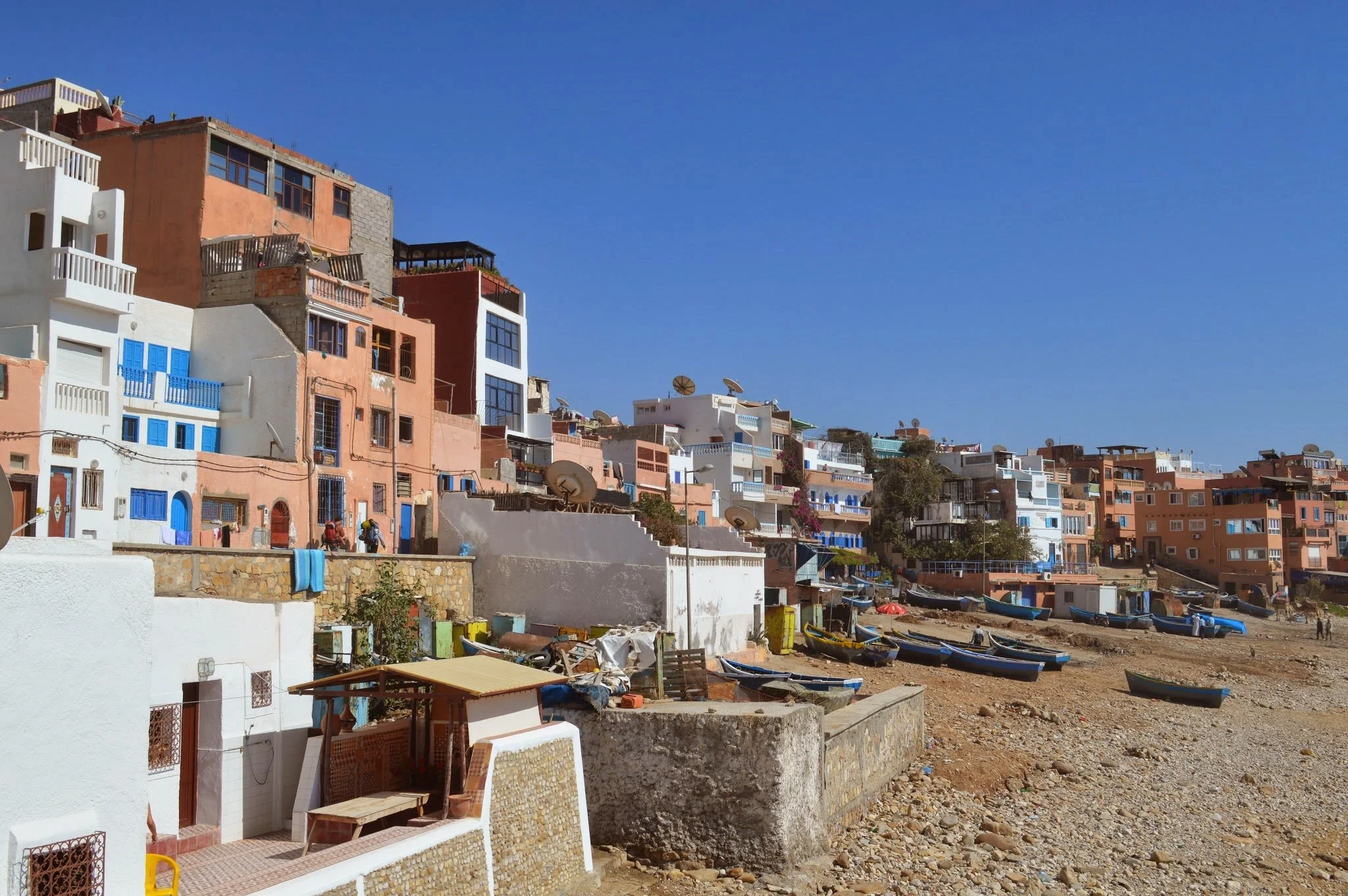 Colorful hillside neighborhood with various multi-story buildings, some with balconies and satellite dishes, located next to a rocky shoreline and boats on the ground, under a clear blue sky.