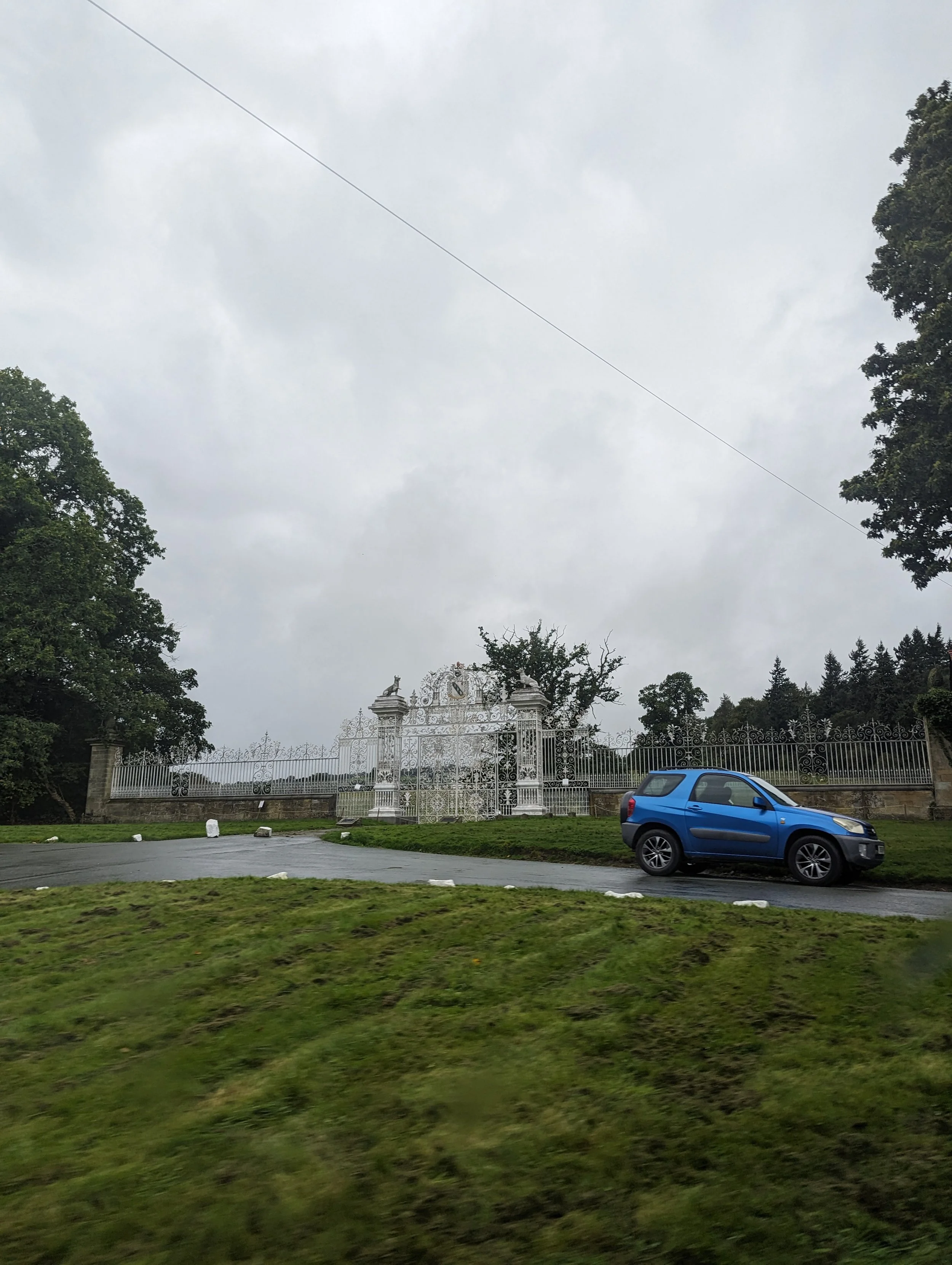 A blue car parked on a wet road in front of a white ornate wrought iron gate and fence, with trees and an overcast sky in the background.