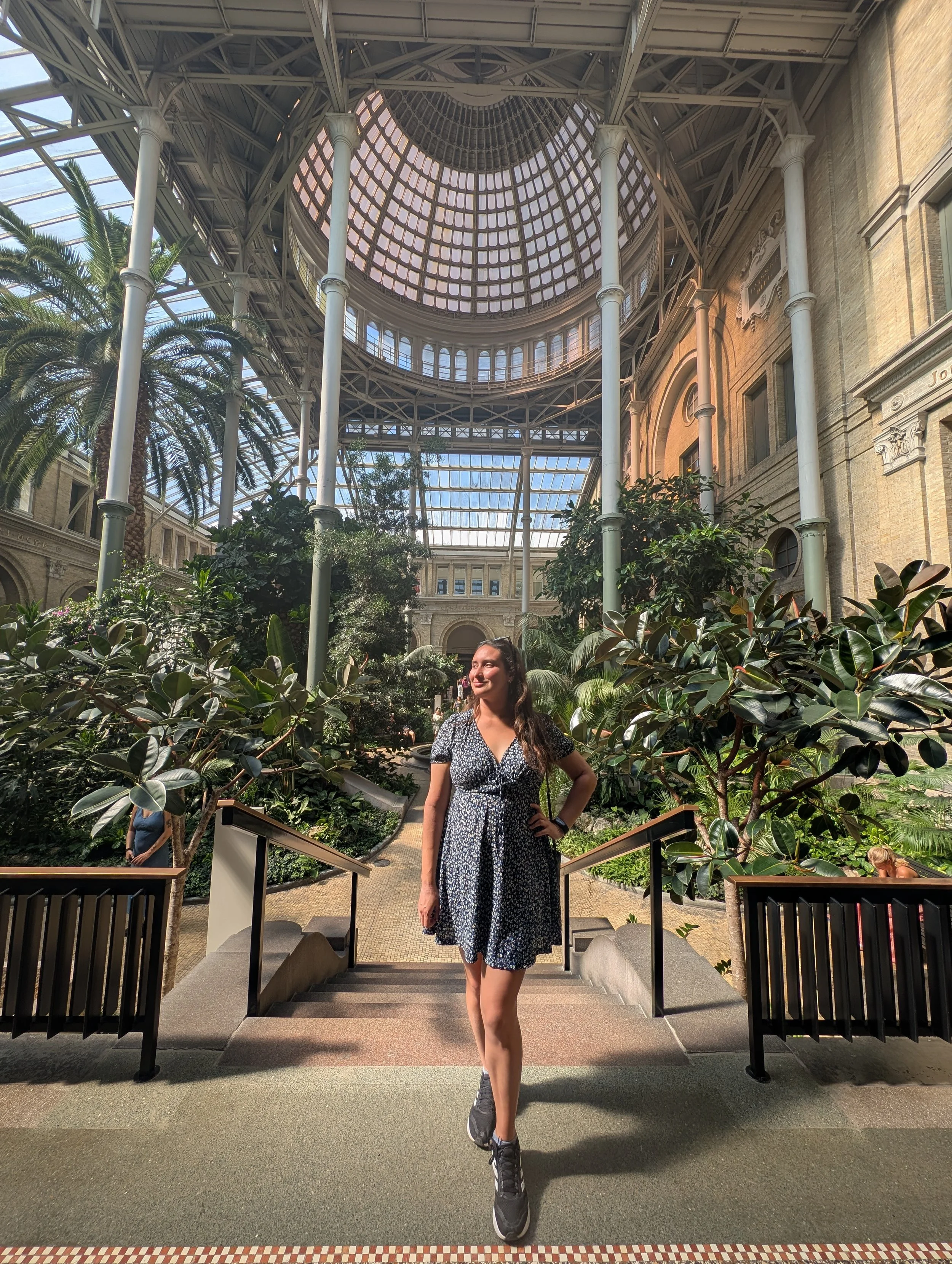 A woman wearing a black and white patterned dress and sneakers standing on stairs inside a building with a glass domed ceiling and surrounded by lush green plants.