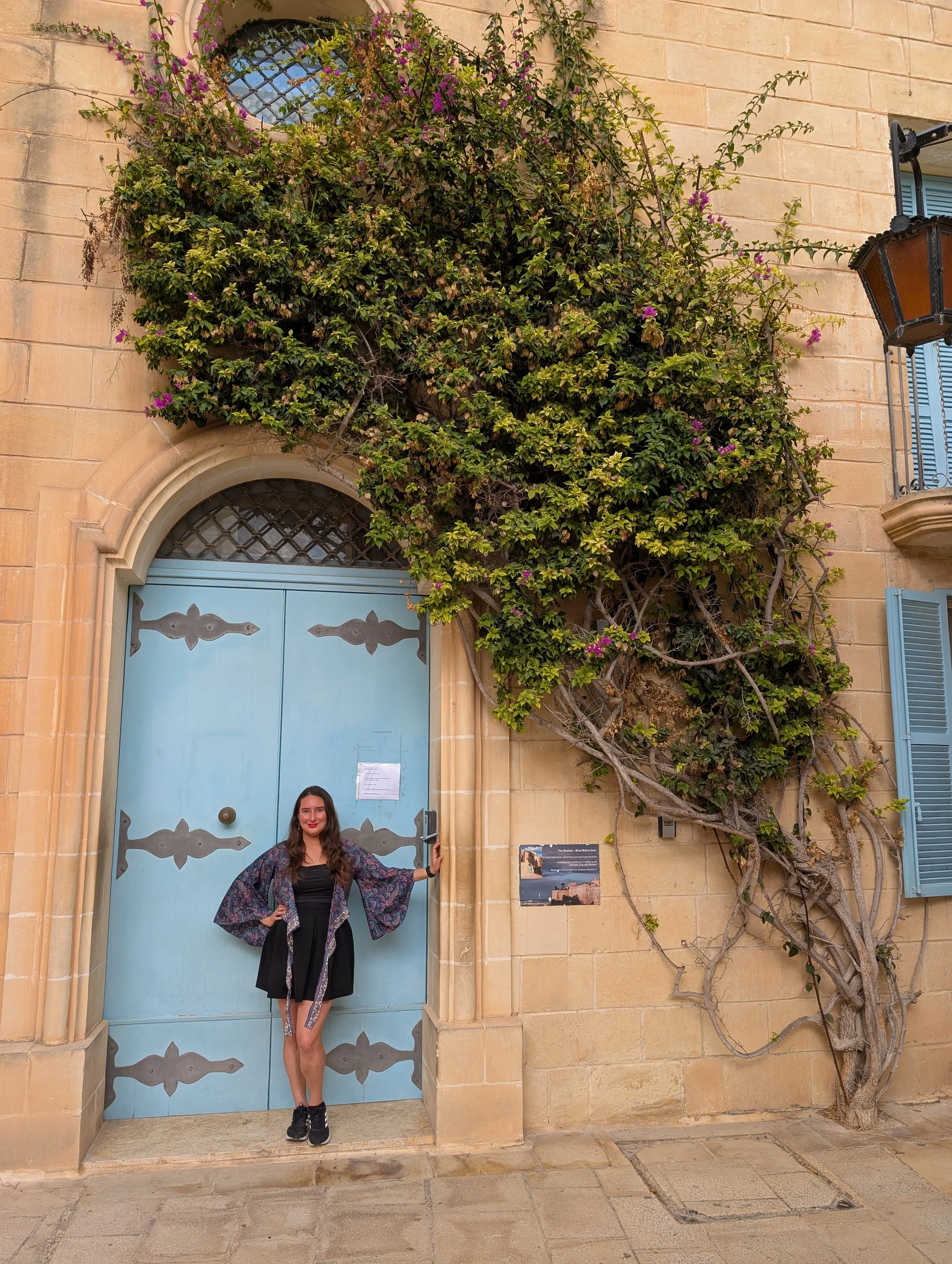 A woman standing in front of a large blue door with decorative patterns, holding the door handle with one hand. The building's exterior is made of beige stone, with a large plant, possibly bougainvillea, growing beside the door, partly covering an ar