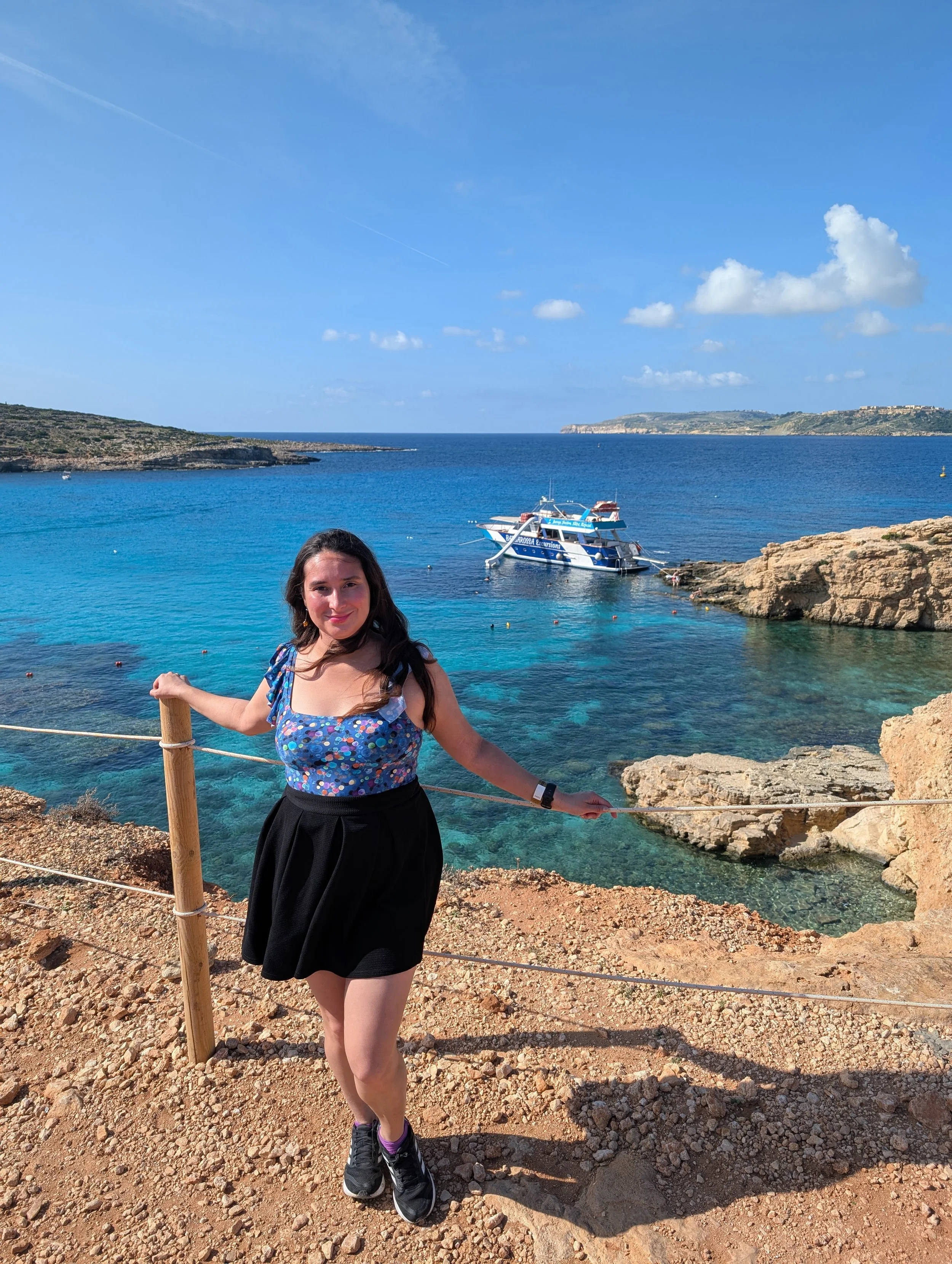 A young woman standing on rocky ground near the coast with clear blue water, rocky cliffs, a boat, and a distant shoreline under a partly cloudy sky.