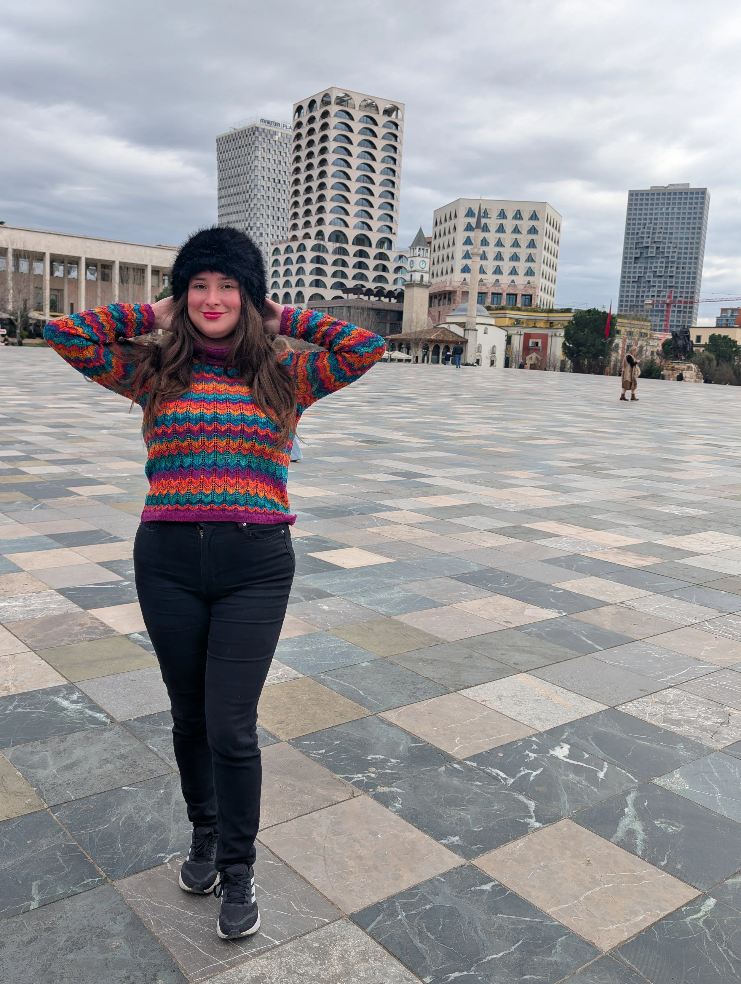 Woman wearing a colourful zigzag-patterned sweater, black pants, black sneakers, and a black furry hat, standing on a large tiled plaza with tall modern buildings in the background under a cloudy sky.