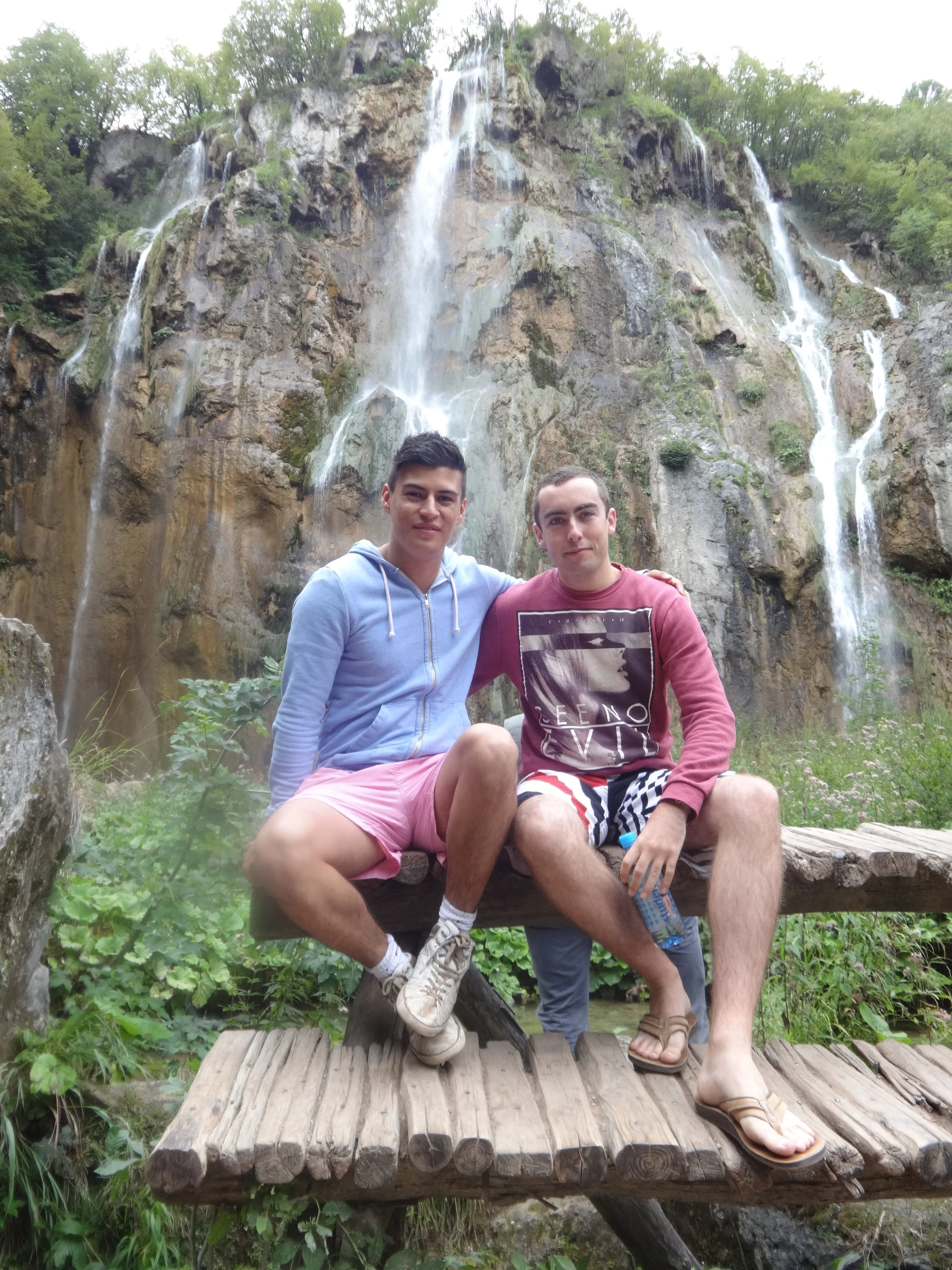 Two young men sitting on a wooden bridge in front of a waterfall surrounded by green trees.