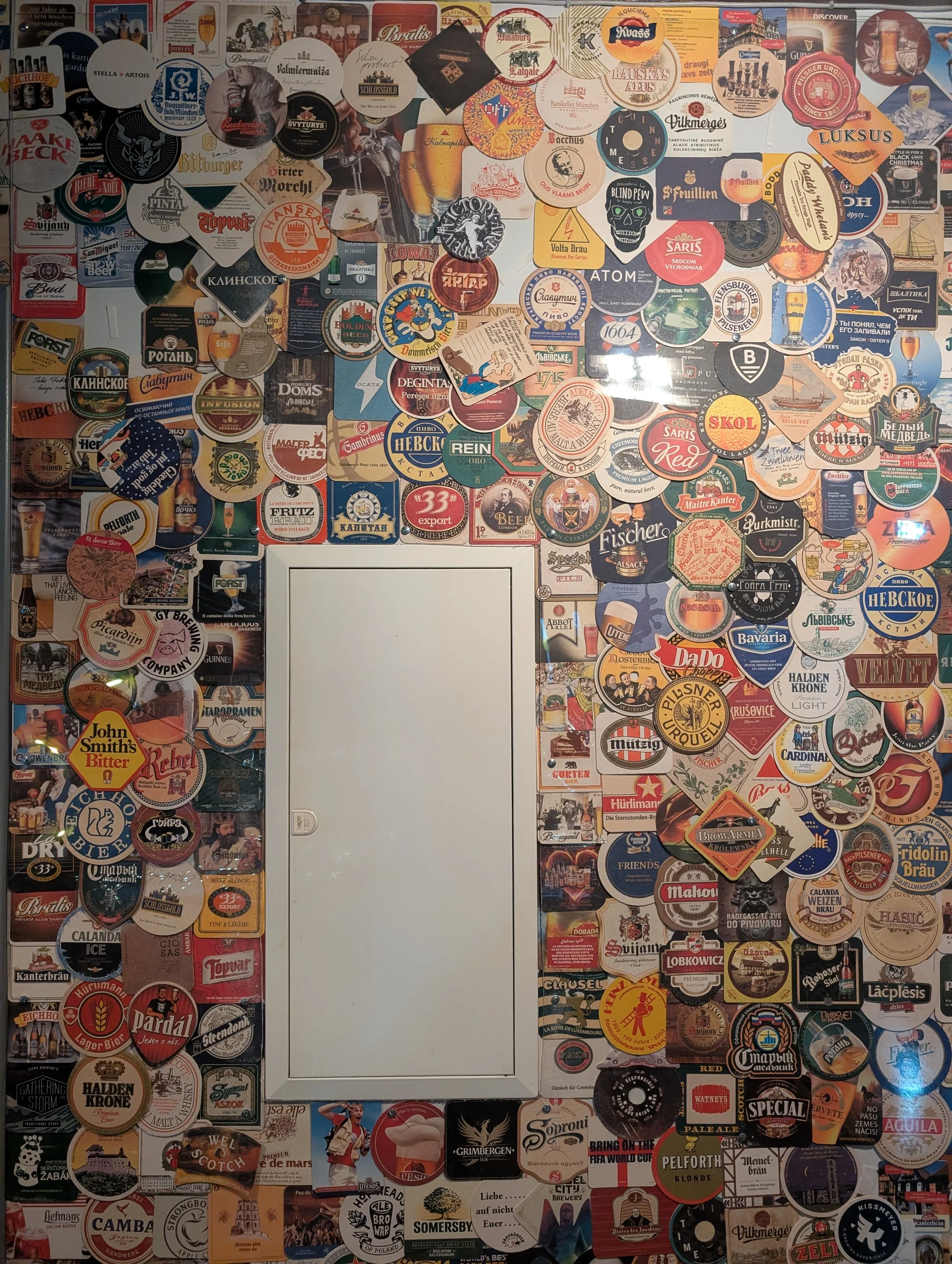 A wall covered with various beer and beverage bottle labels and stickers surrounding a white door or panel.