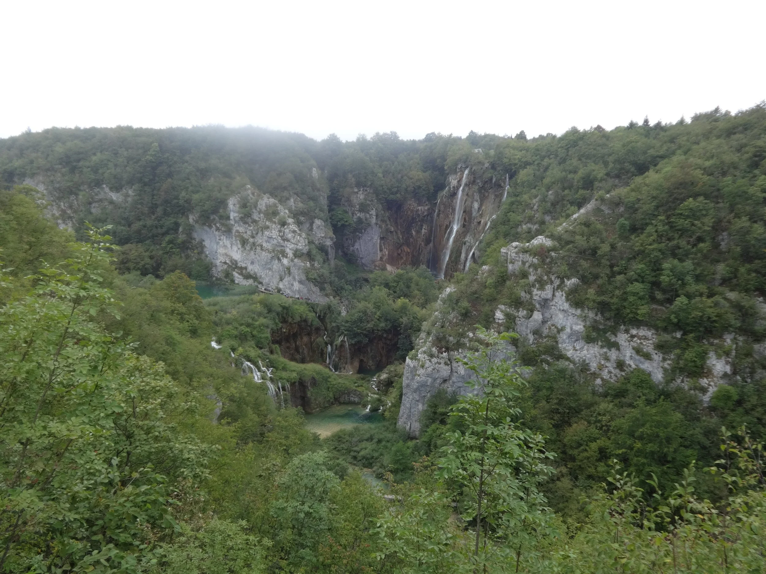 A scenic view of a lush green valley with waterfalls cascading down rocky cliffs, surrounded by dense forest under a foggy sky.