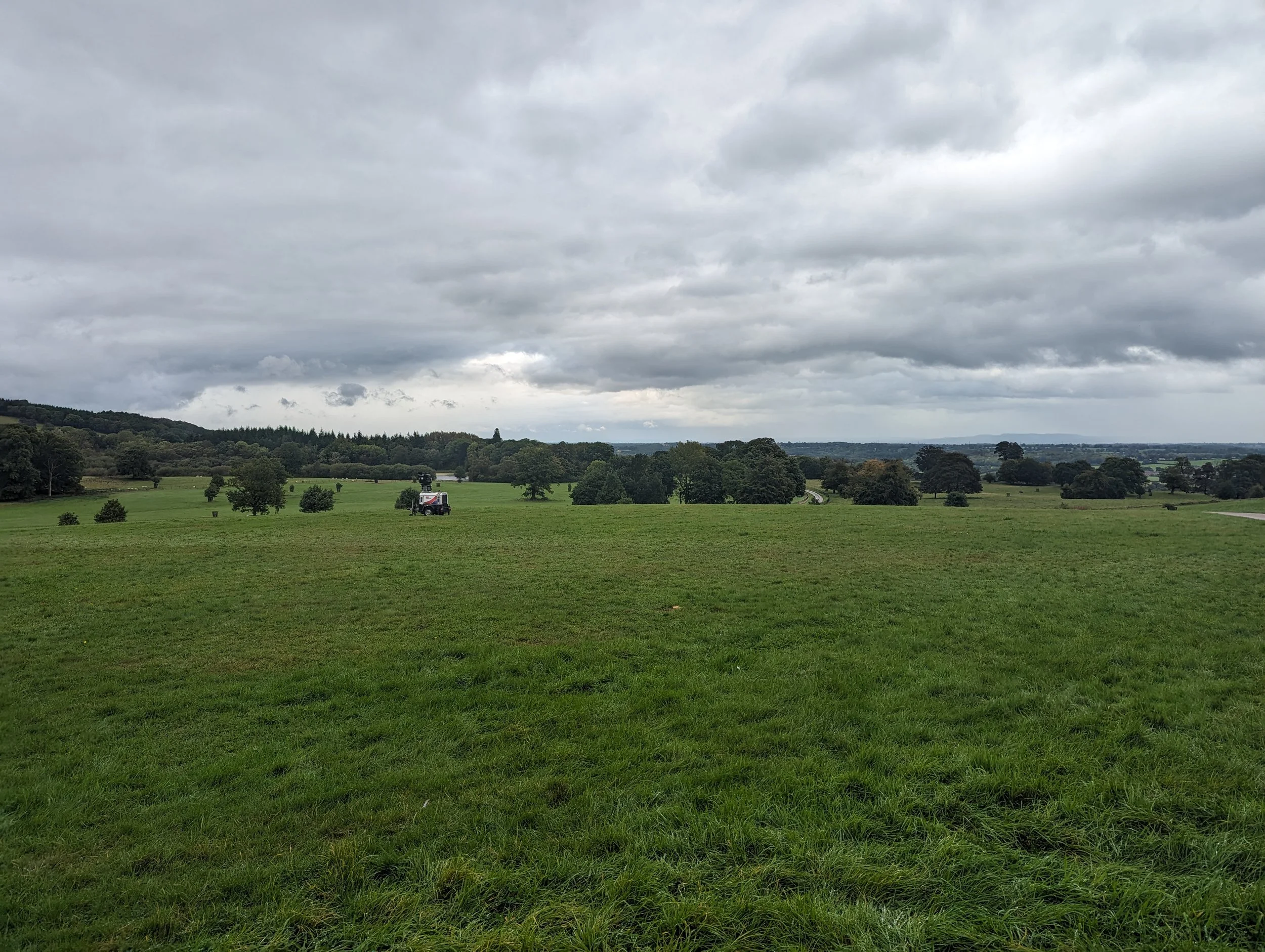 A wide green field with small trees and a wet weather vehicle captures overcast clouded sky.