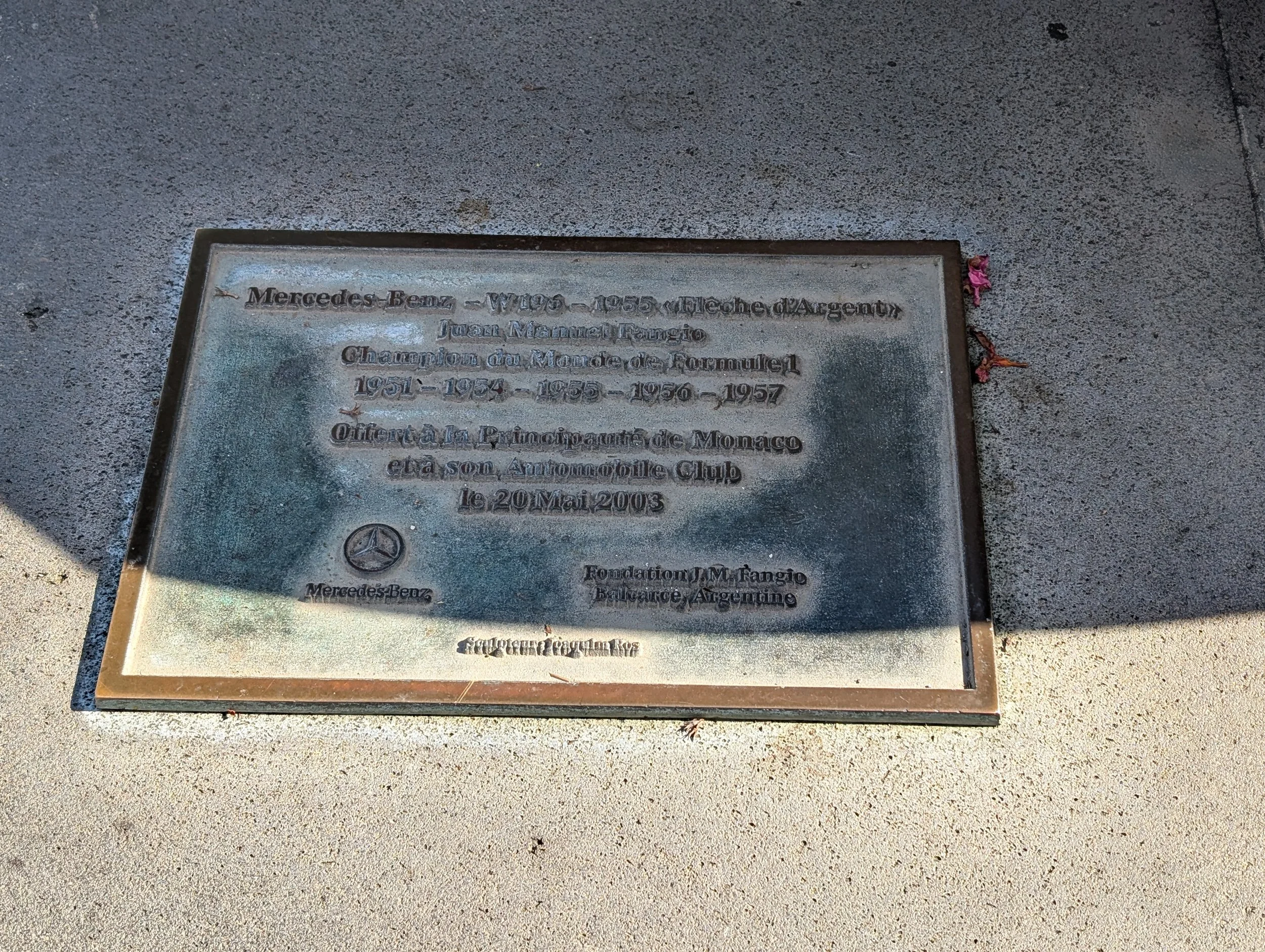 A metal plaque on the sidewalk celebrating Mercedes Benz's racing achievements from 1951 to 1957, with a Mercedes Benz logo at the bottom.