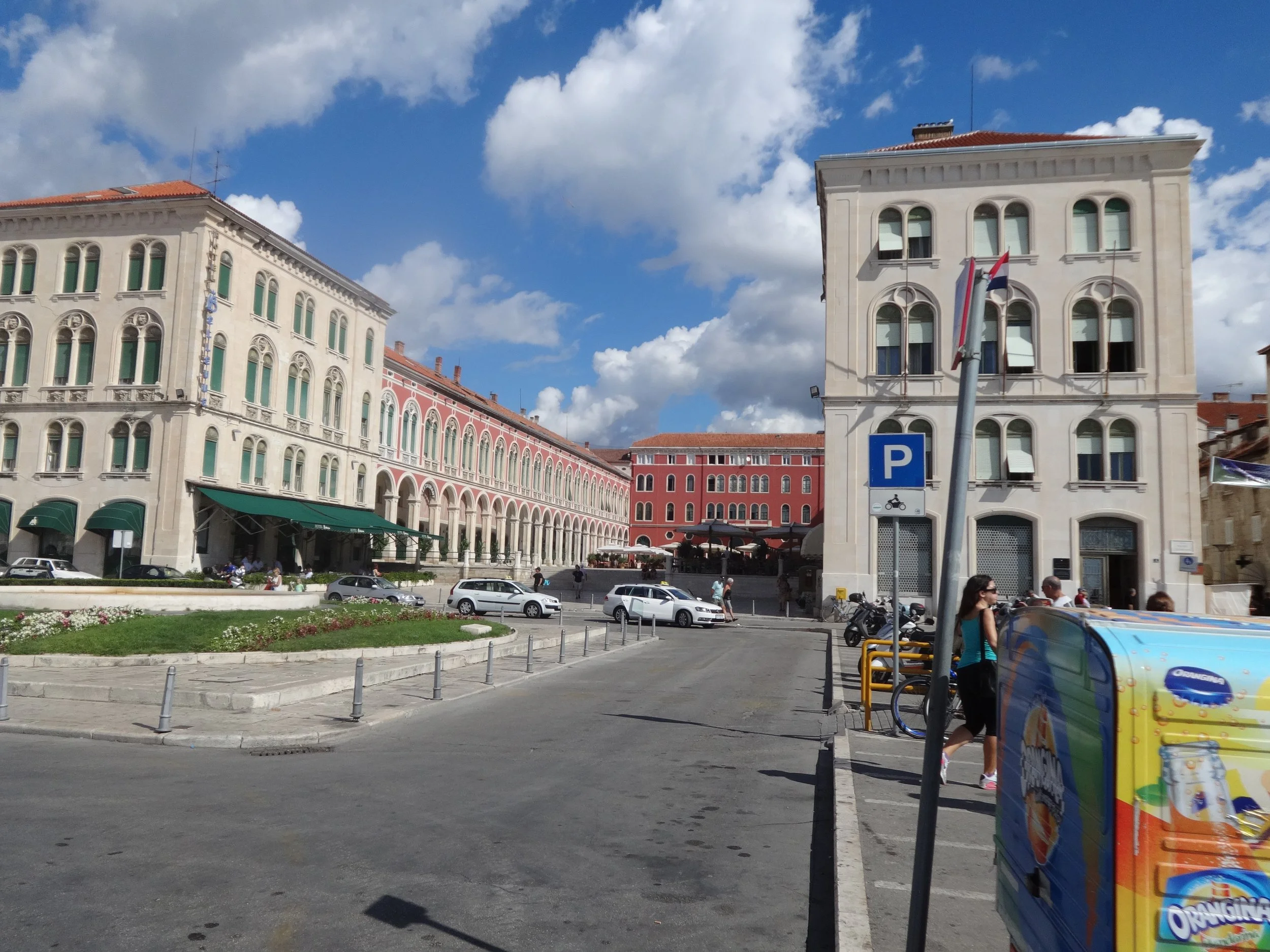A busy city square with historic European-style buildings, parked cars, pedestrians, and a vending machine in the foreground. The sky is partly cloudy.