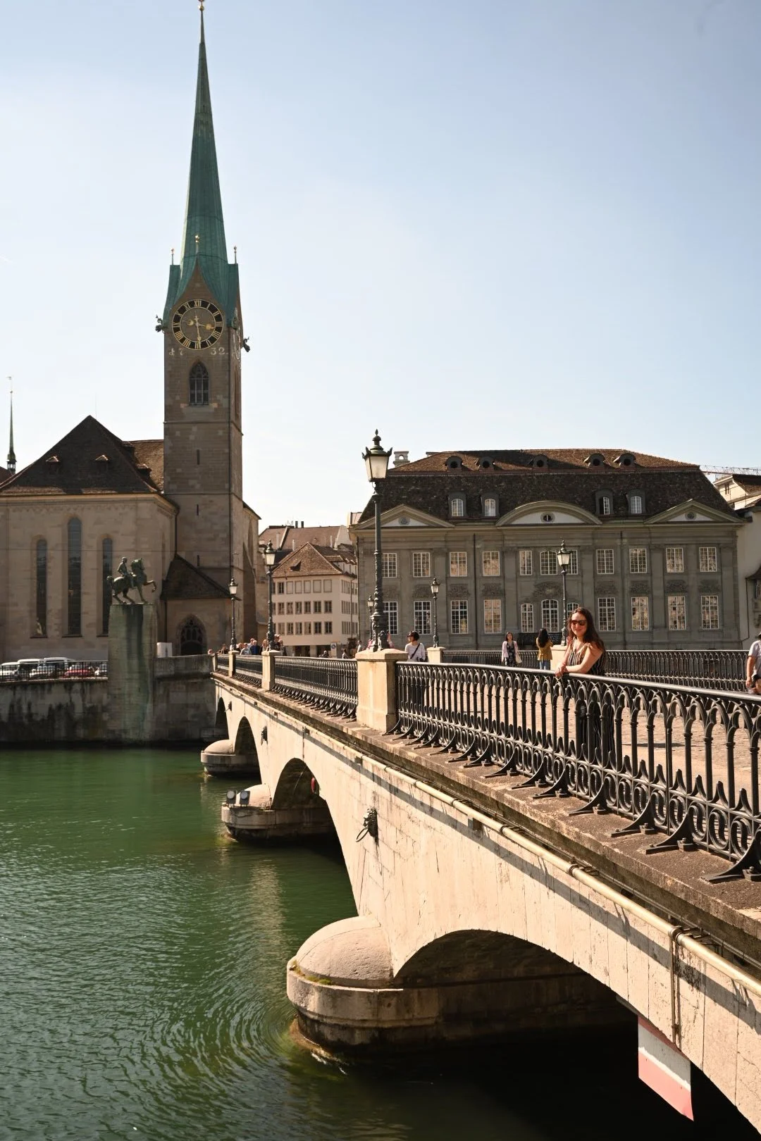 A historic European cityscape with a church featuring a tall clock tower and a green spire, a stone bridge crossing a green river, and people walking along the bridge on a sunny day.